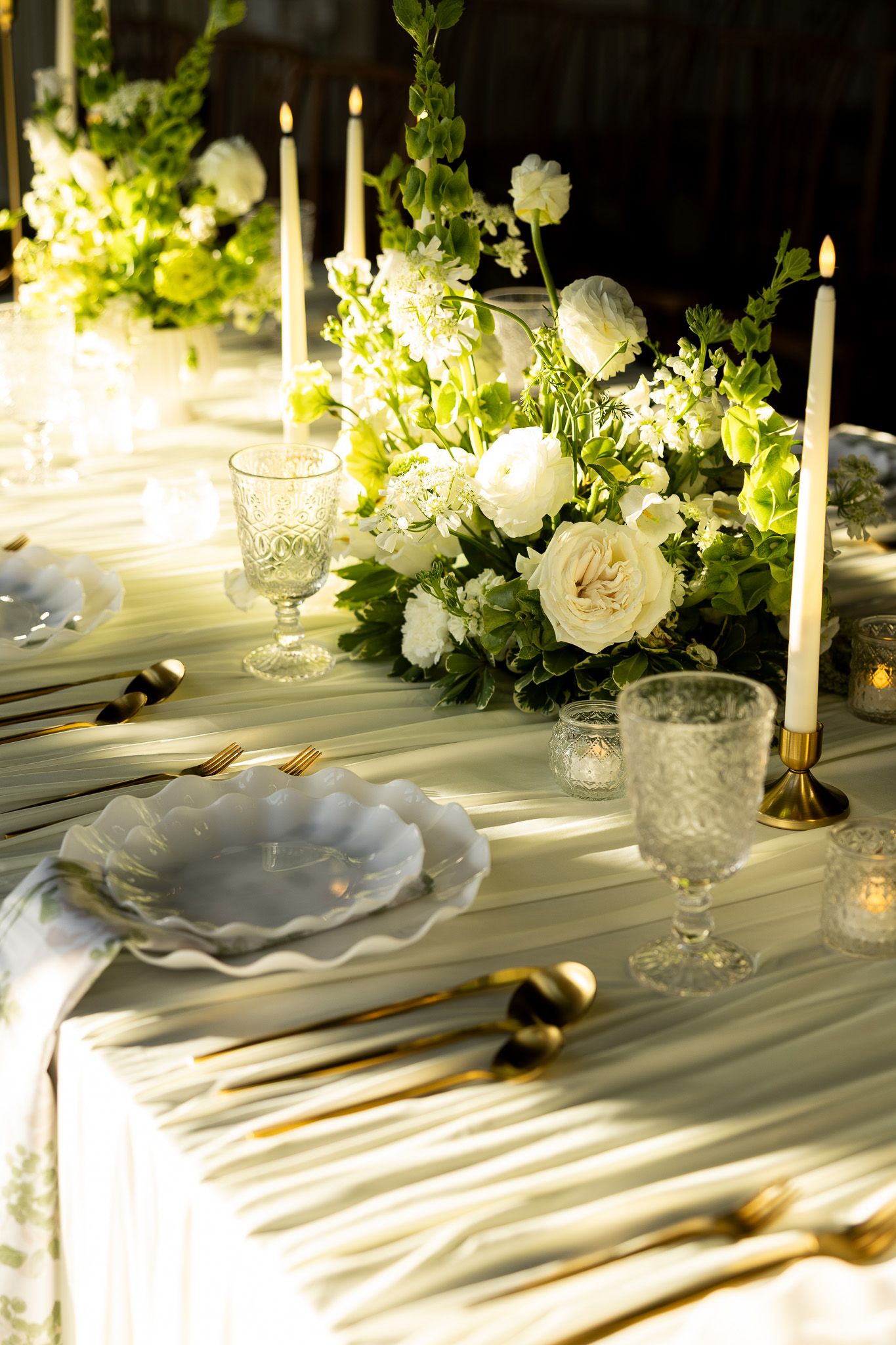 Elegant wedding table setting with white roses, greenery, gold cutlery, crystal glasses, and tall candles on a white linen tablecloth.