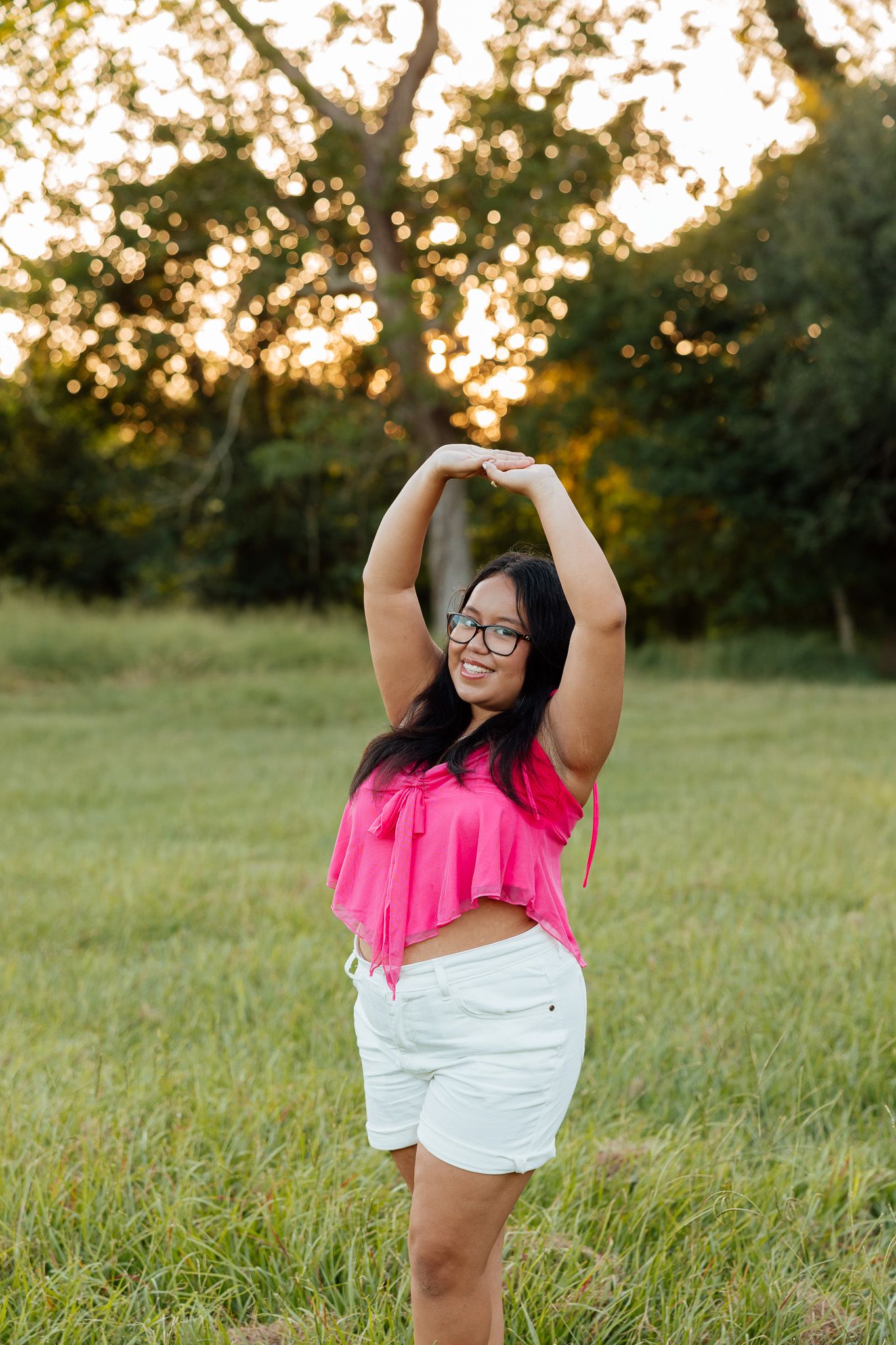 A young woman in a pink crop top and white shorts poses with arms raised in a sunny green field at golden hour.