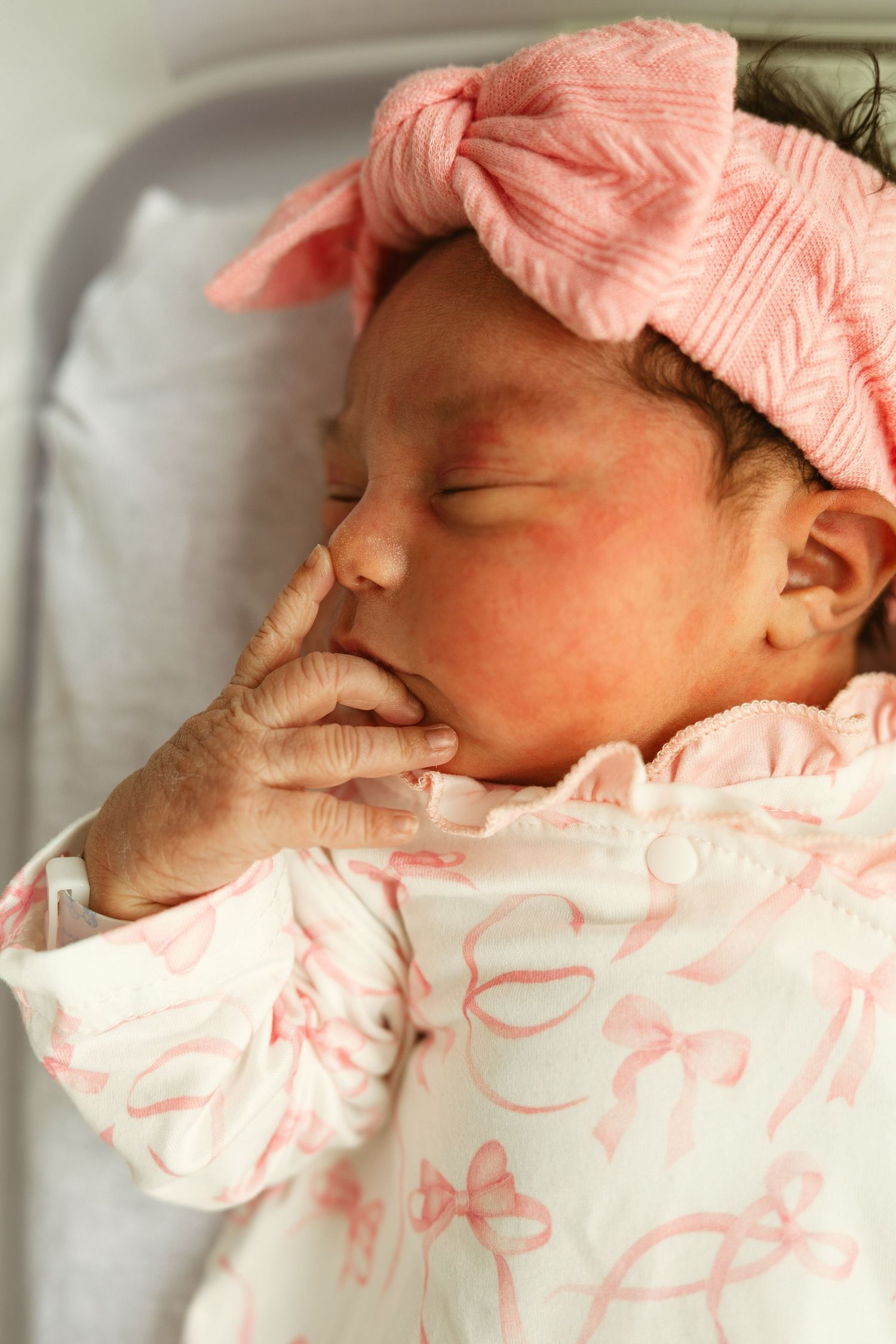 Sleeping newborn baby girl wearing a pink bow headband and white onesie with pink bows, resting peacefully in hospital bassinet.