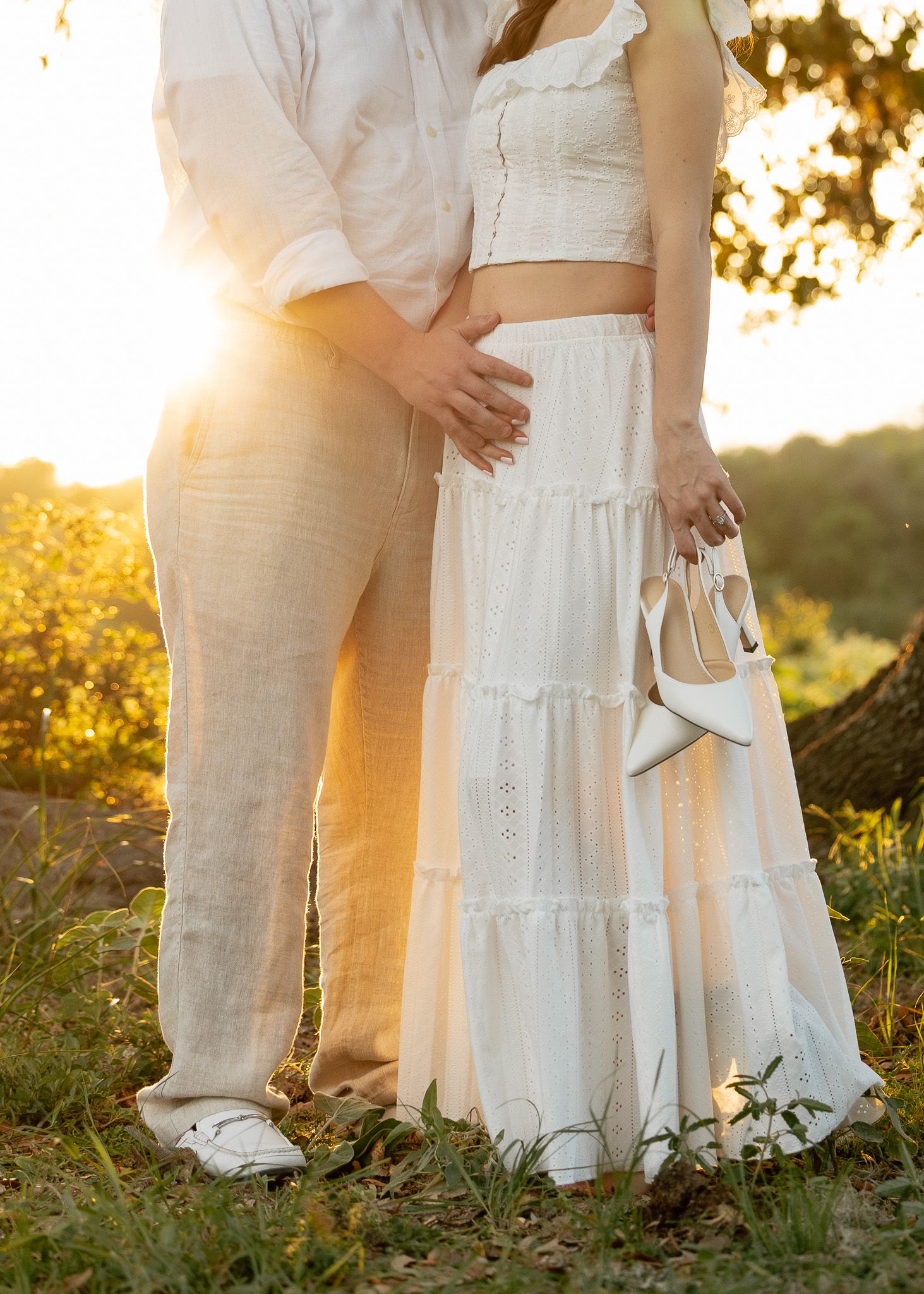 Couple in white outfits embracing at sunset, woman holding heels while partner touches her baby bump outdoors.