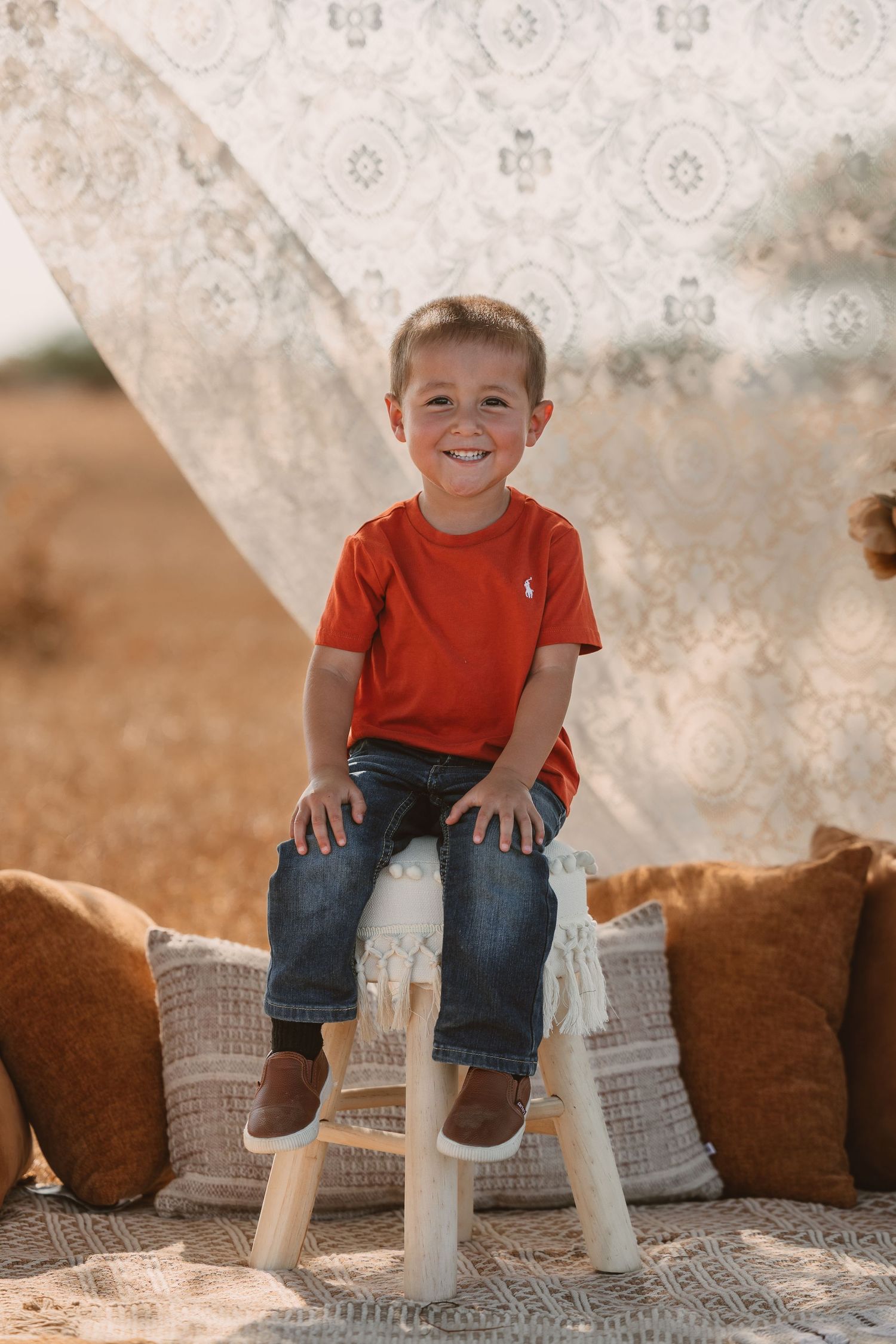 A smiling young boy in a red shirt and jeans sits on a white stool outdoors with boho pillows and lace backdrop.