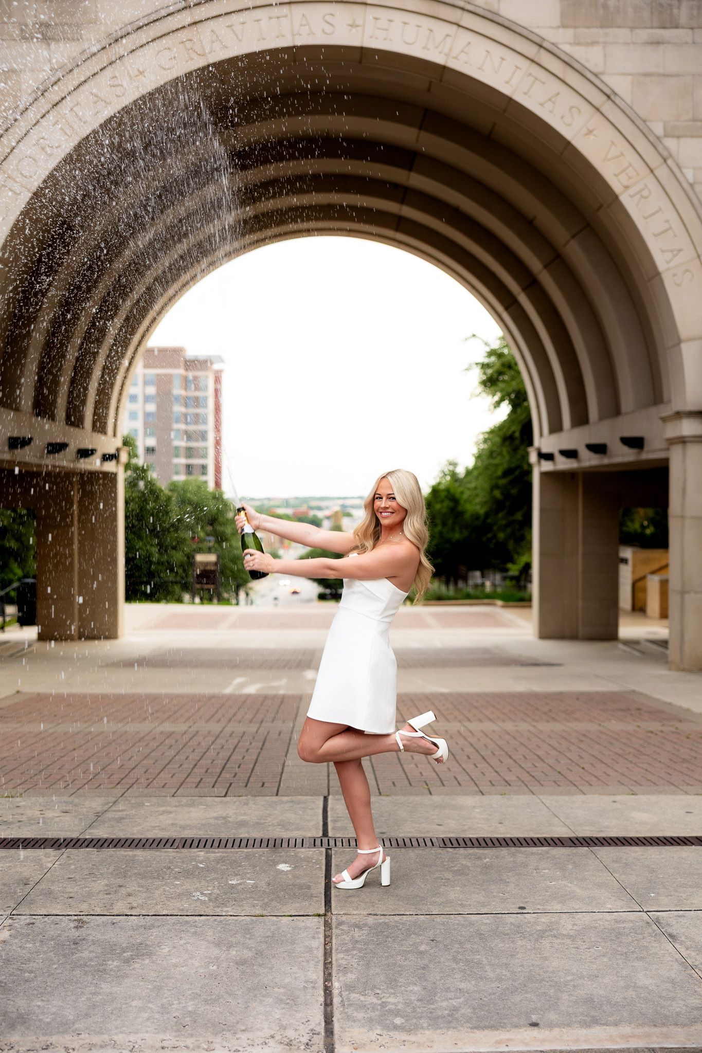Joyful blonde woman in white mini dress popping champagne under large stone archway, posing on one foot.