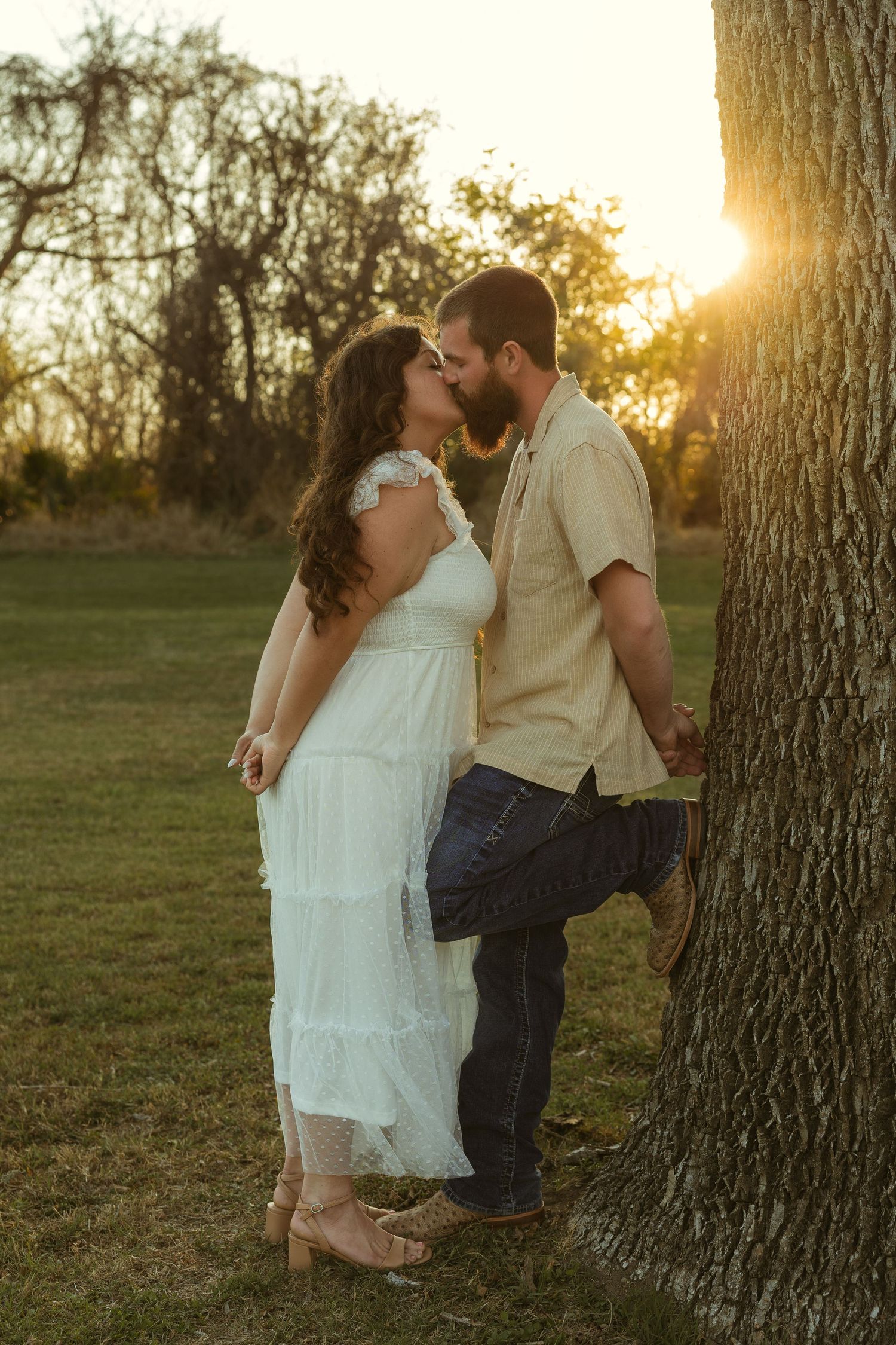 Couple sharing a romantic kiss near a tree at golden sunset in a park, woman in white dress, man in casual attire.
