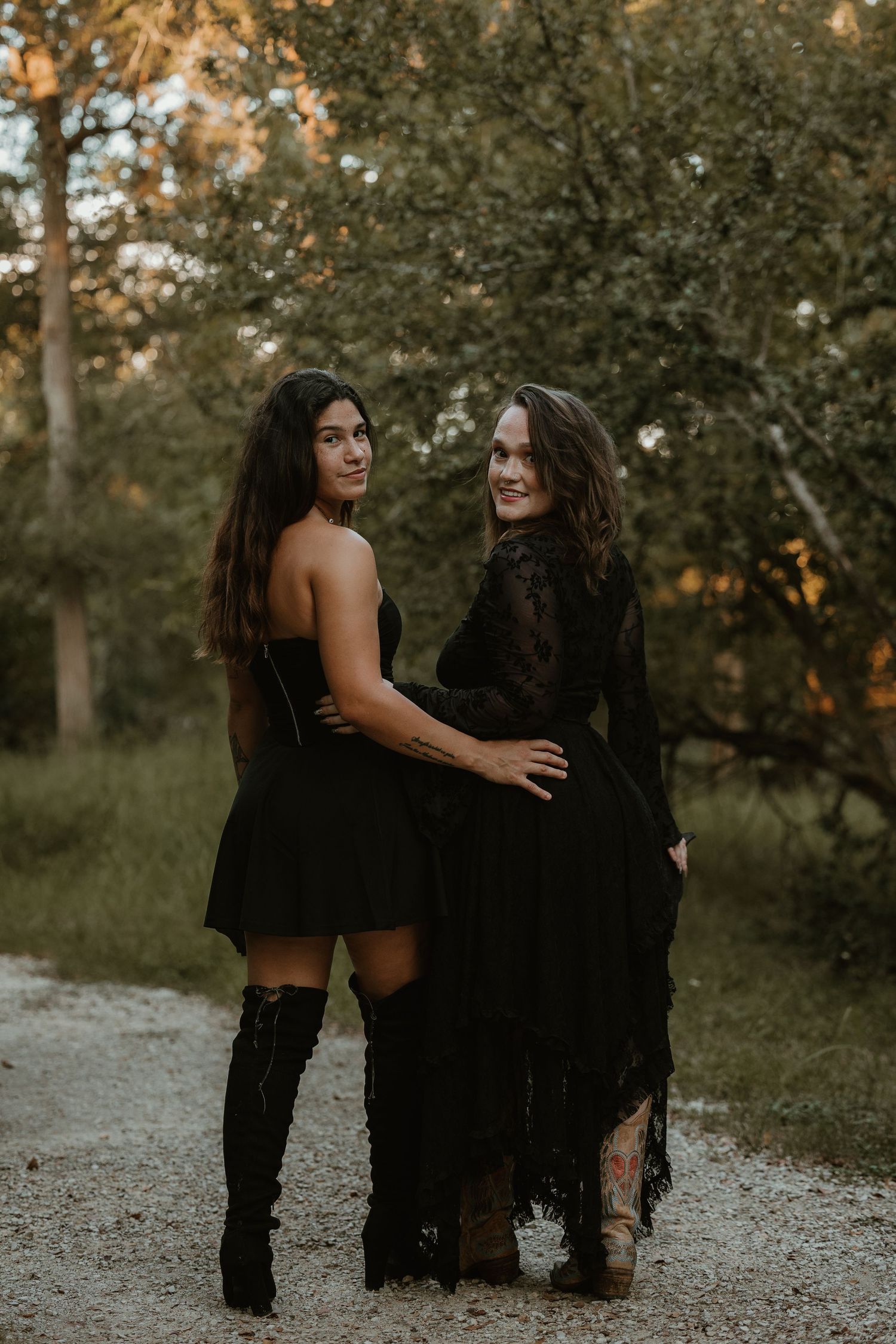 Two friends in black dresses pose together on nature trail at sunset.