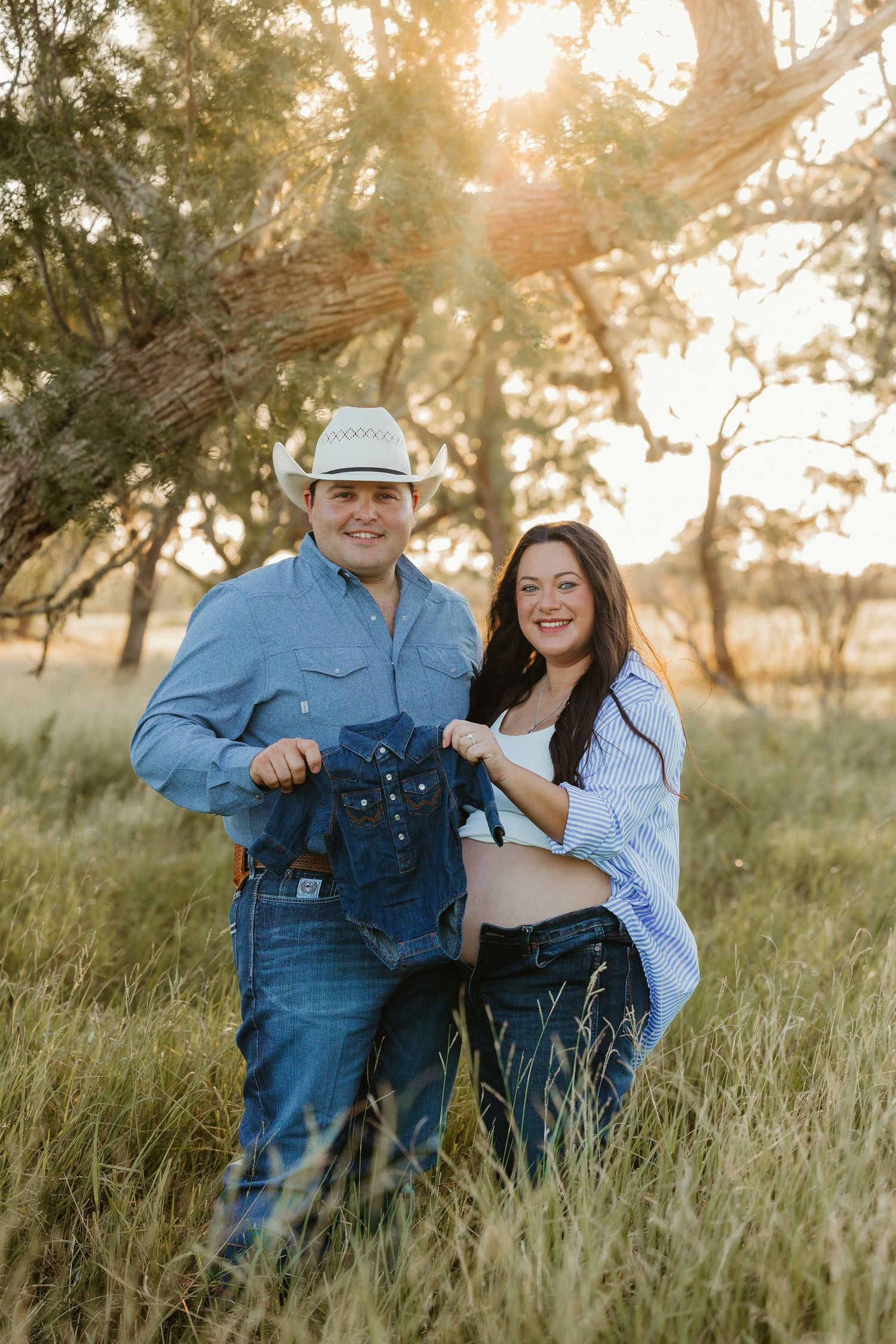 A couple in western attire poses together in a grassy field at sunset.