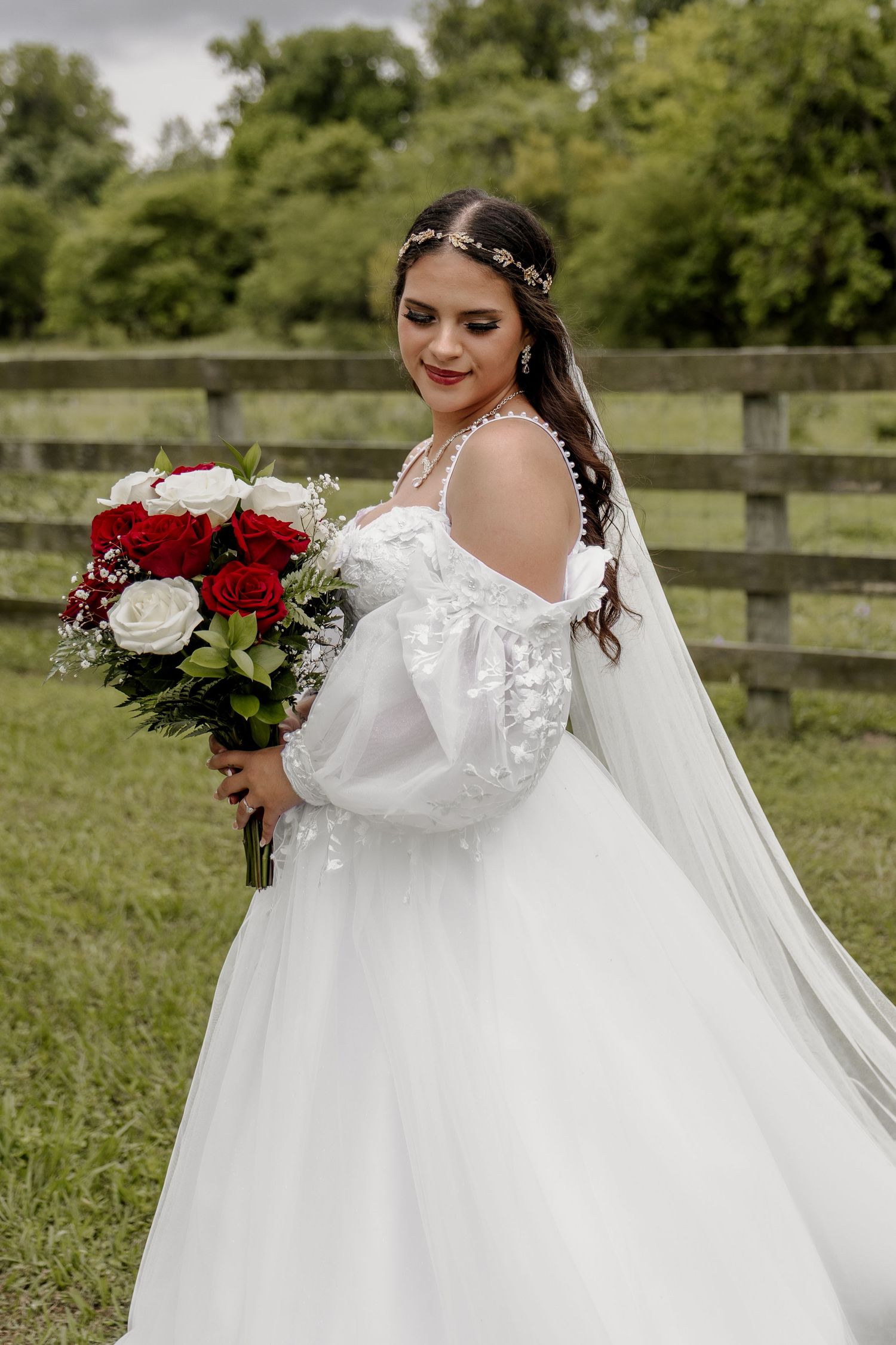 Bride in flowing white gown holding red and white bouquet against rustic fence.