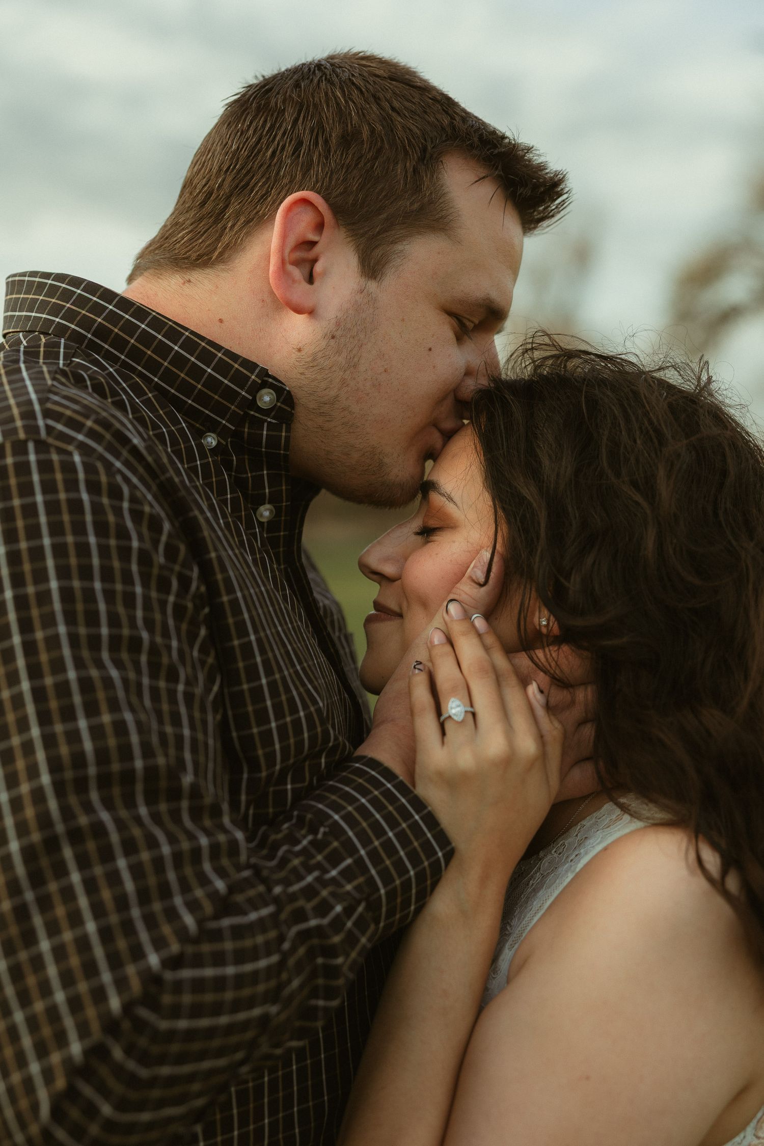 Romantic couple sharing a tender kiss outdoors, woman displaying an engagement ring while man gently holds her face.