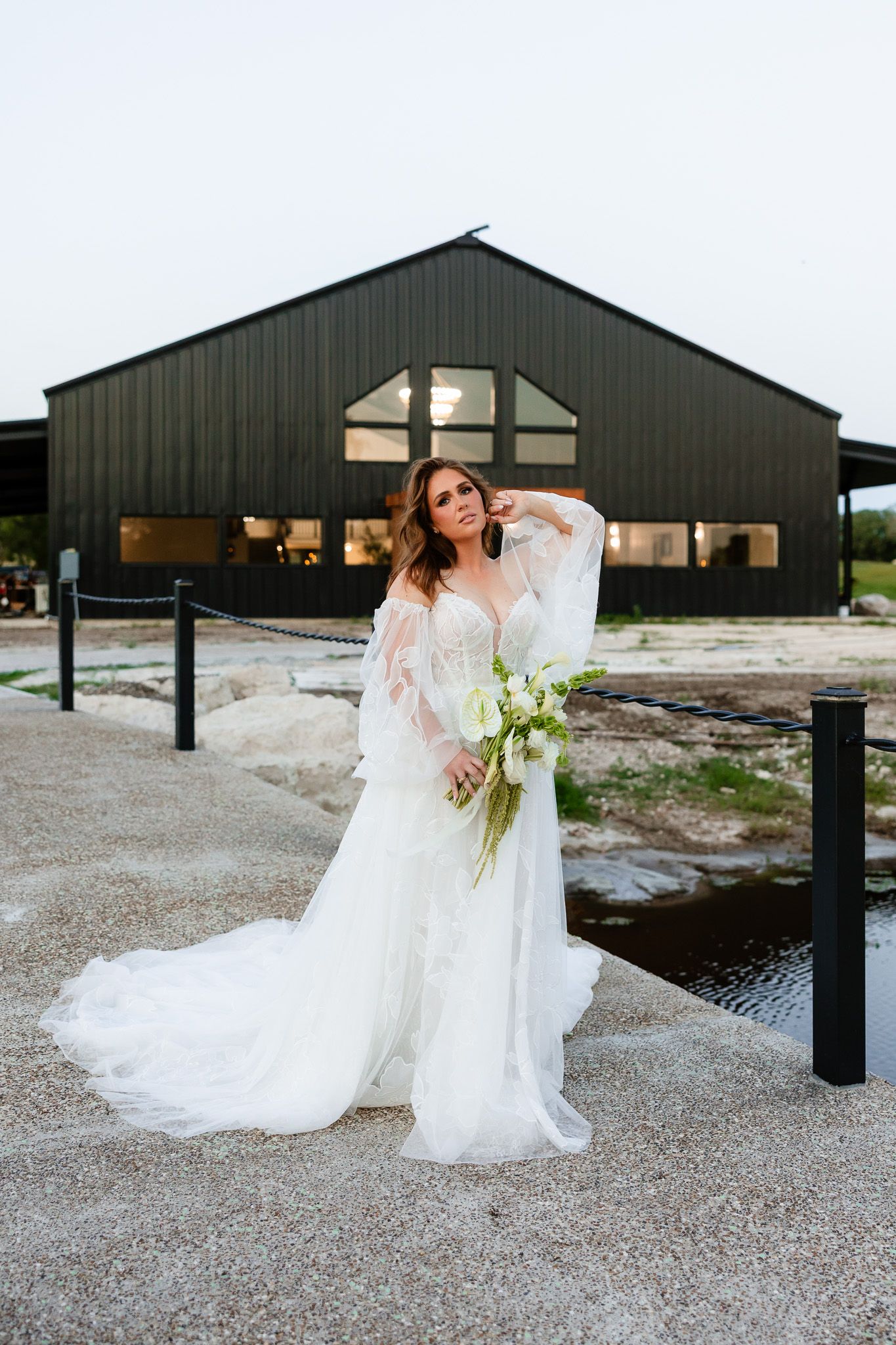 Bride in a flowing white tulle gown holds a white bouquet, posing before a modern black barn venue.