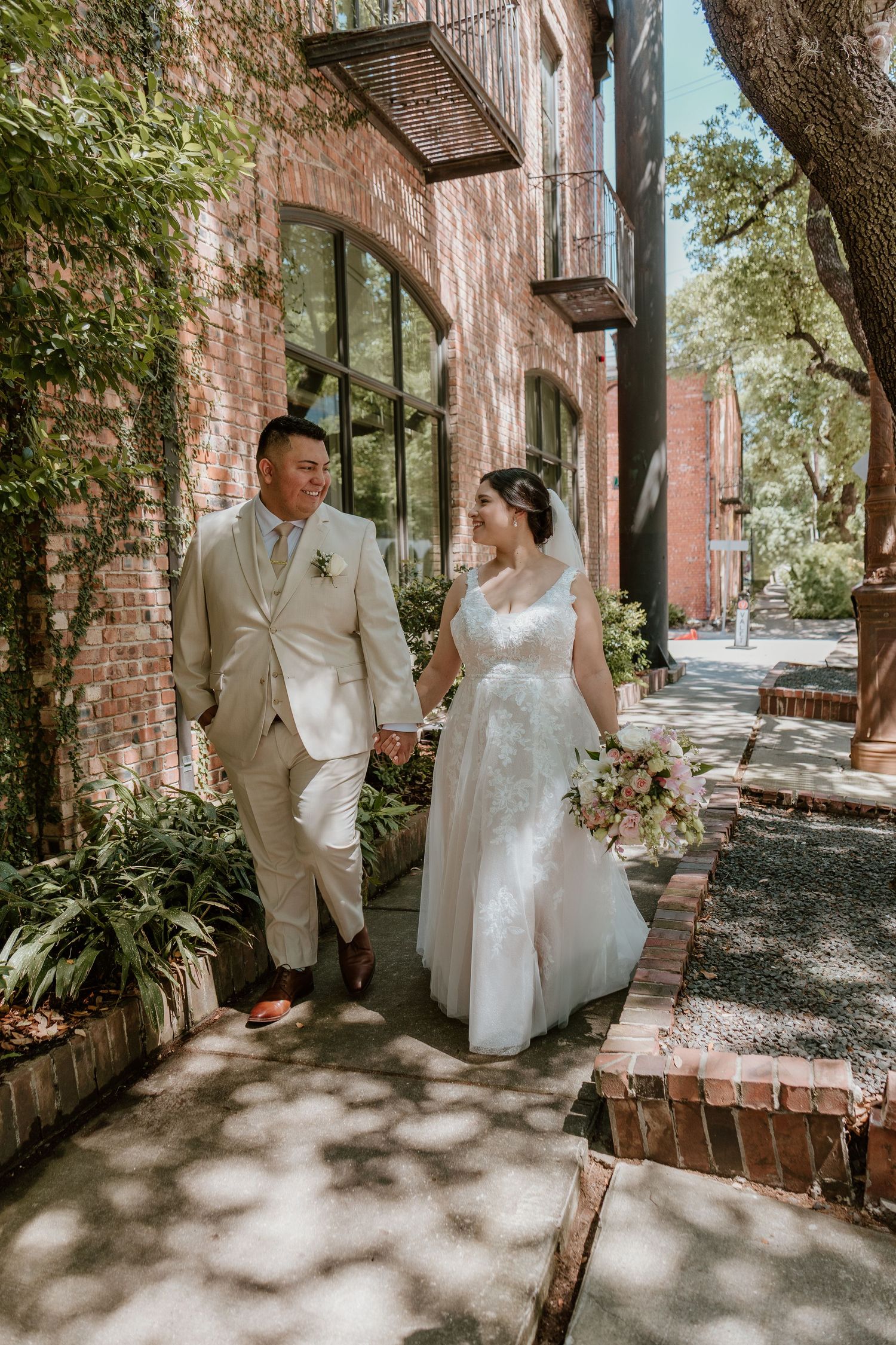 A couple in wedding attire walks along a brick building sidewalk on their special day.
