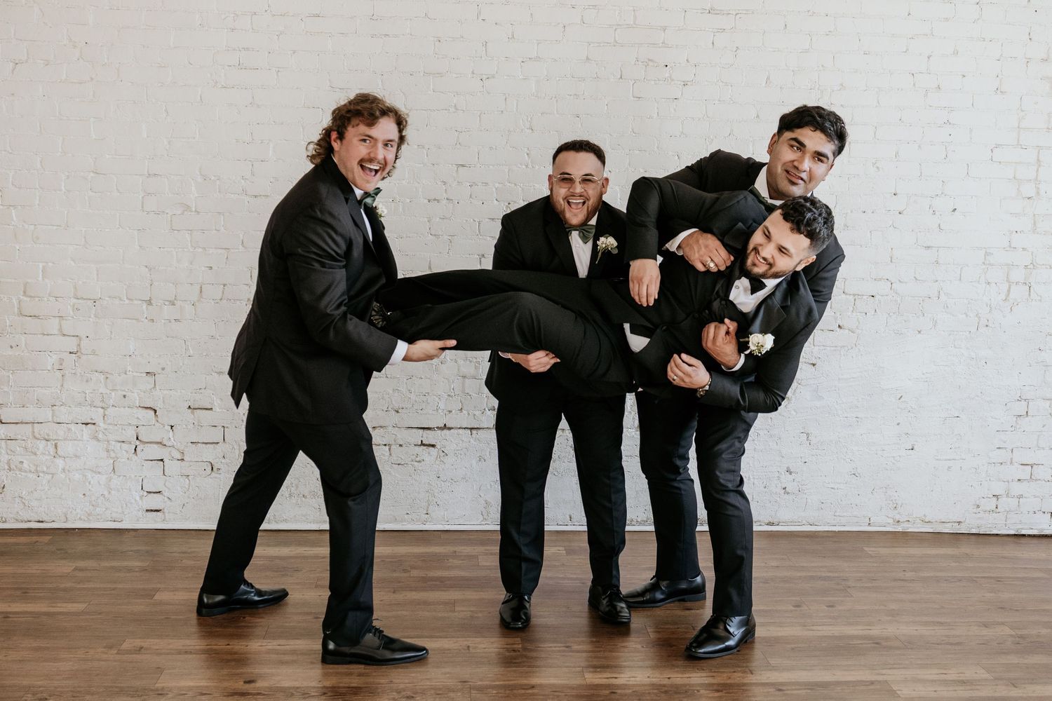 Groomsmen in black suits performing a fun pose against a white wall.