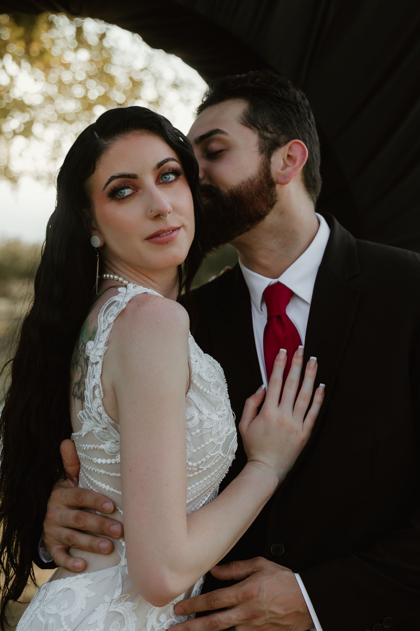 A romantic wedding couple embraces outdoors with the bride in a white lace dress and groom wearing a red tie.