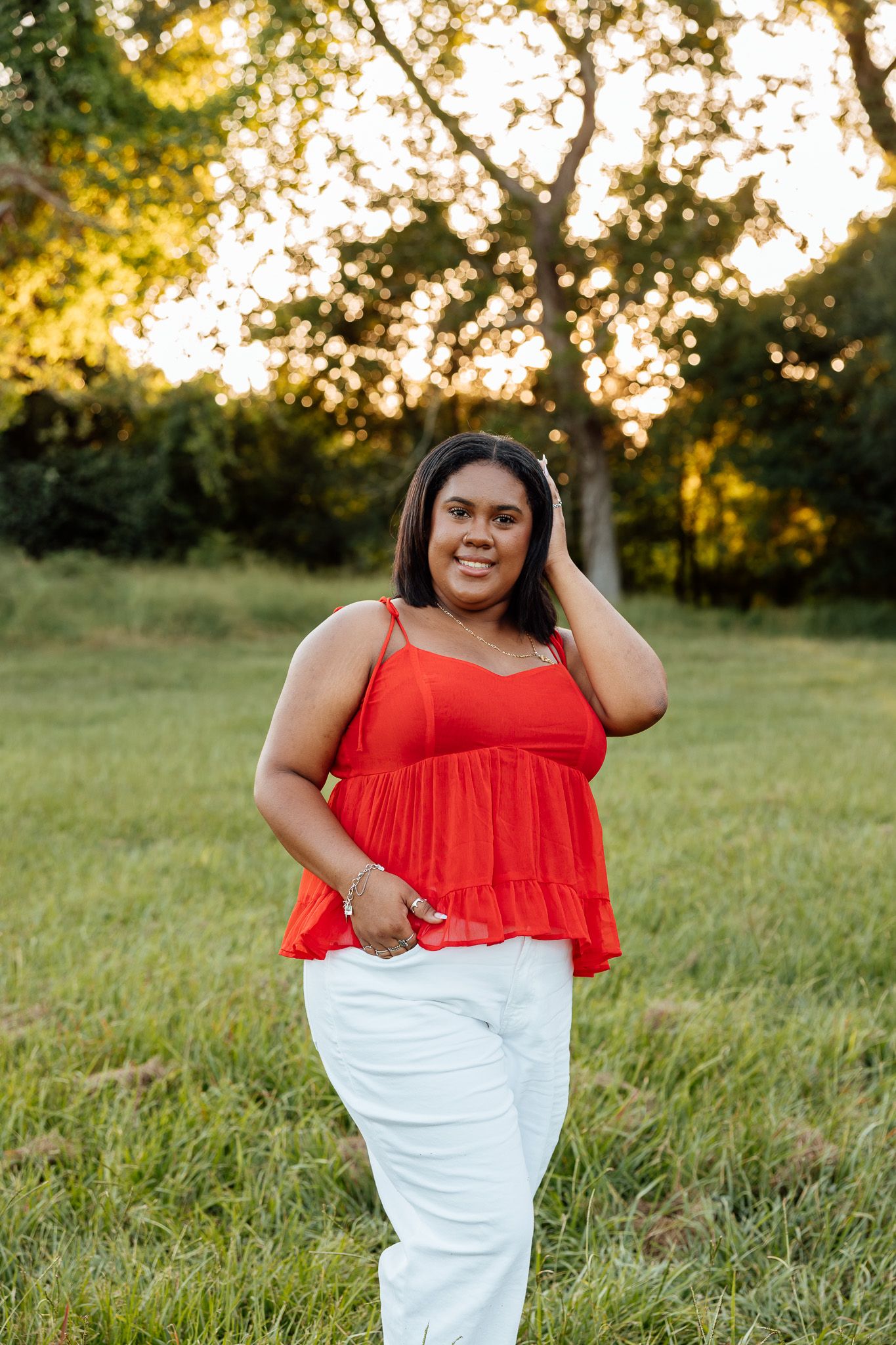 Woman in red top and white pants smiling outdoors at golden hour with green trees in background.