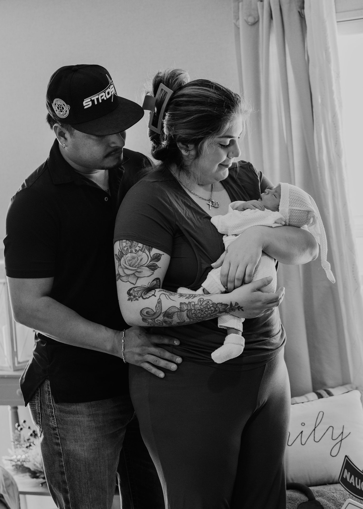 Black and white photo of new parents lovingly gazing at their newborn baby, mother with floral arm tattoo.