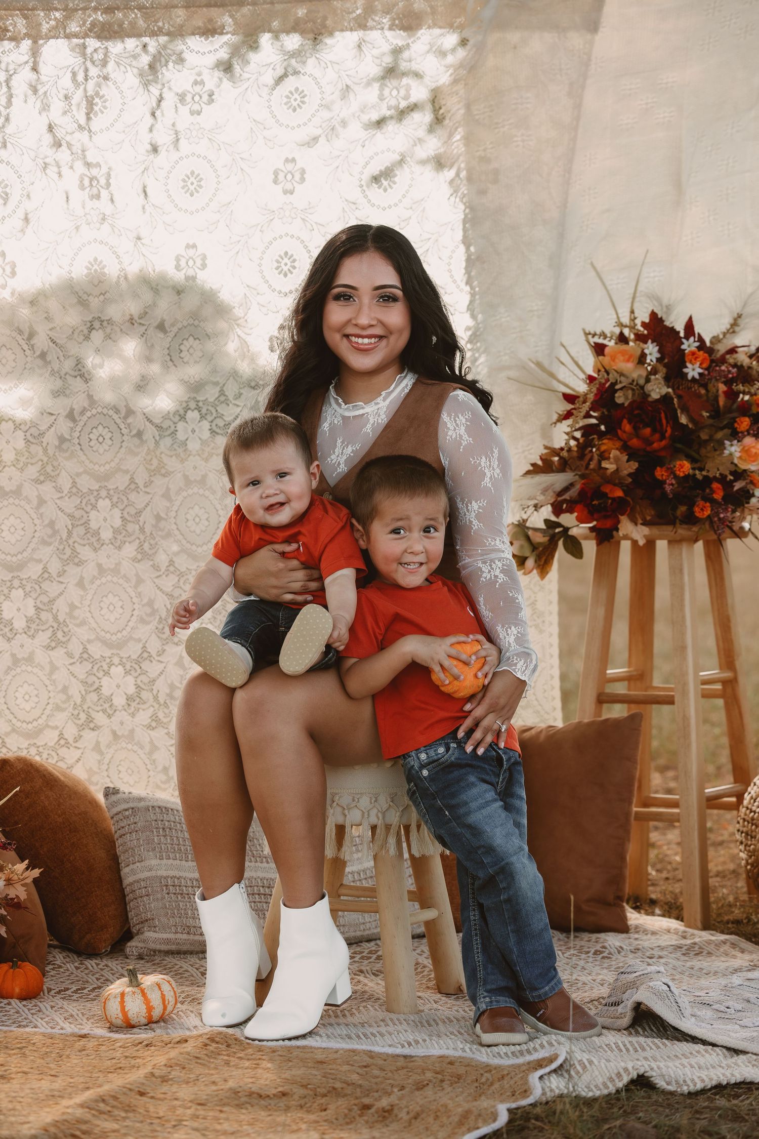 A smiling woman sits with two young boys in orange shirts, surrounded by fall decor and pumpkins in a warm autumn photo session.