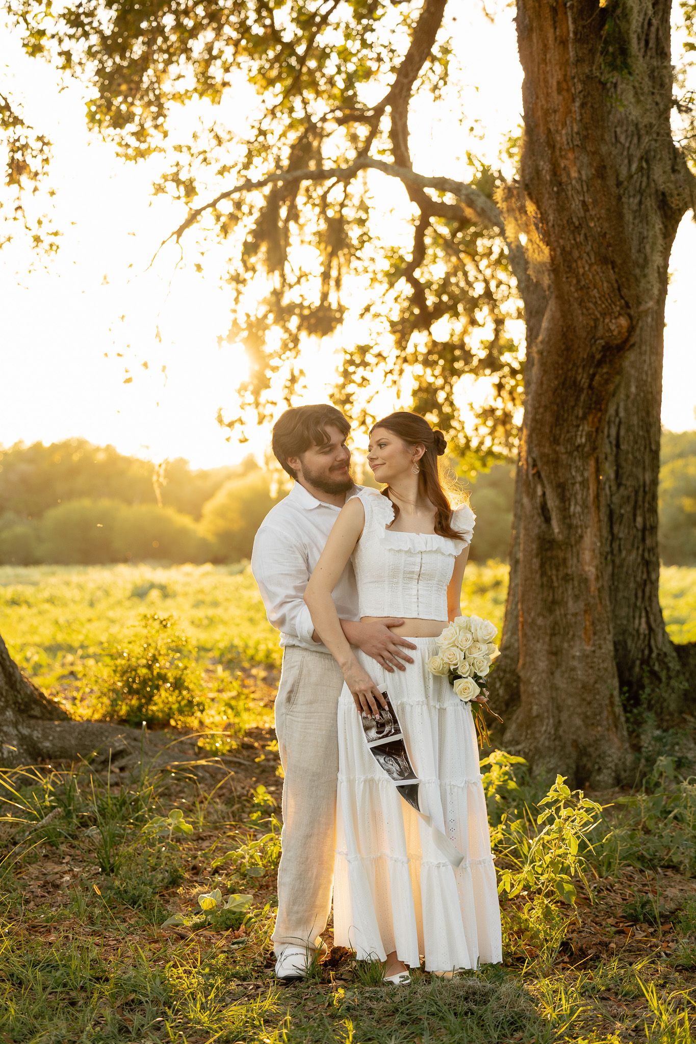 Couple in white outfits embracing under oak tree at golden sunset in a green field, woman holding ultrasound photos.