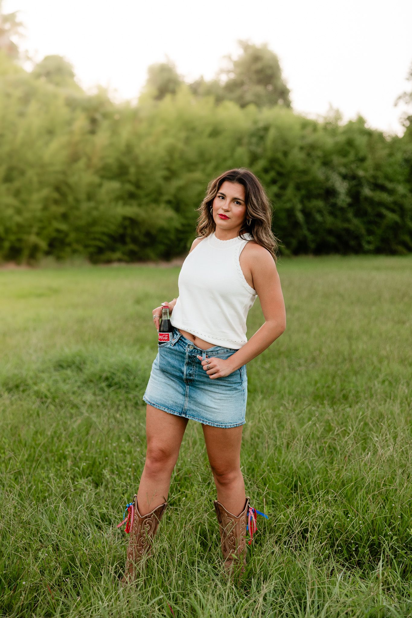 Young woman in white crop top, denim mini skirt, and cowboy boots standing in a green field.