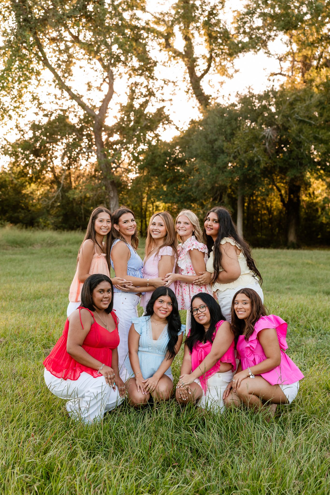 Group of nine young women posing outdoors in a sunny park, wearing colorful pink and white outfits.