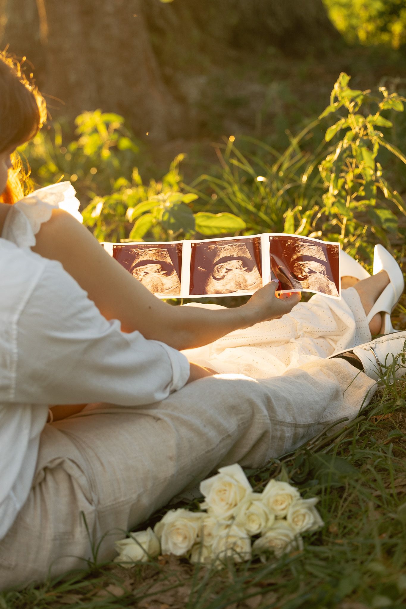 Pregnant woman sitting outdoors in golden sunlight holding ultrasound photos beside white roses bouquet.