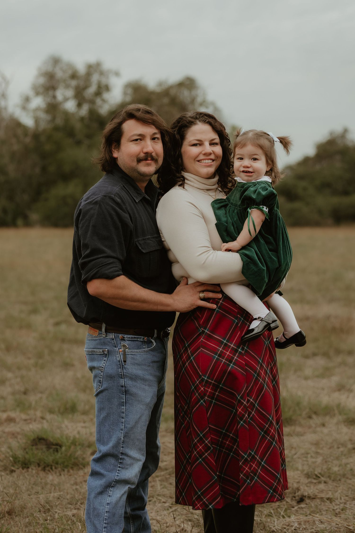Family portrait of parents and toddler girl in green dress posing in a field during fall season.