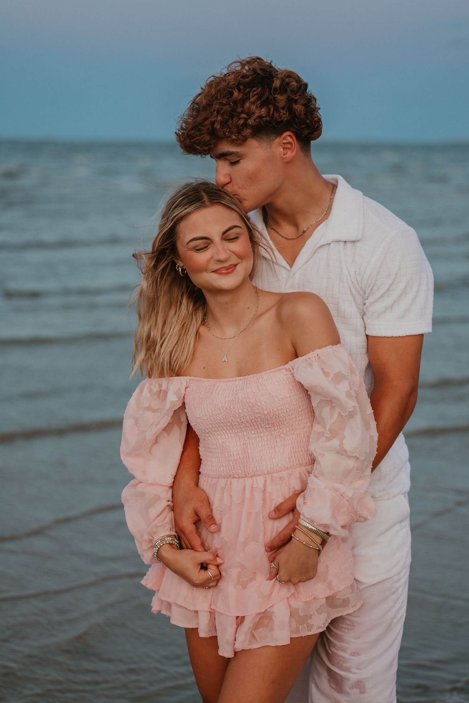 A romantic beach photoshoot featuring a couple in pink and white outfits against the ocean.