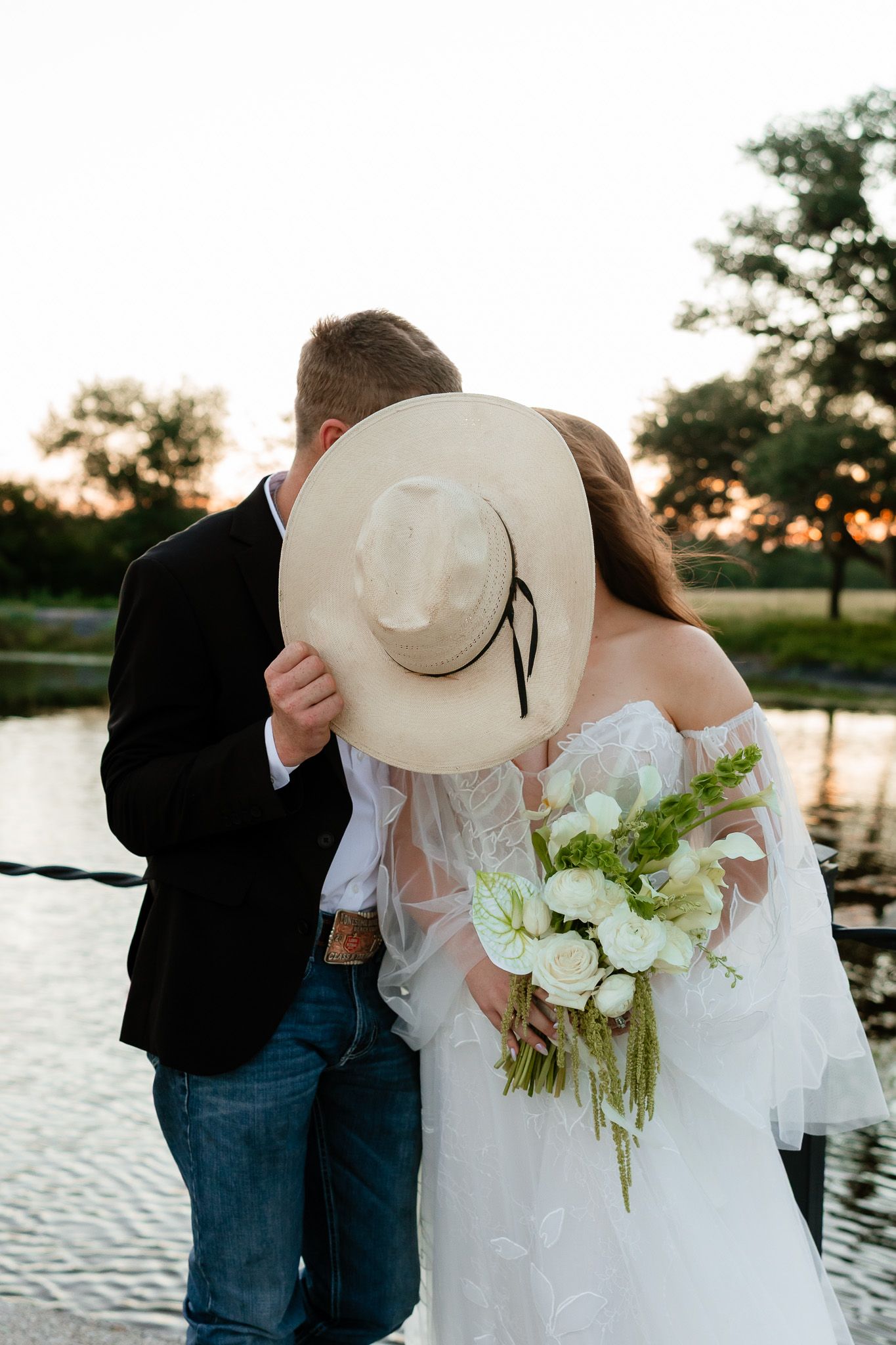 Bride and groom kissing behind a white cowboy hat at sunset near a pond, bride holding white floral bouquet.