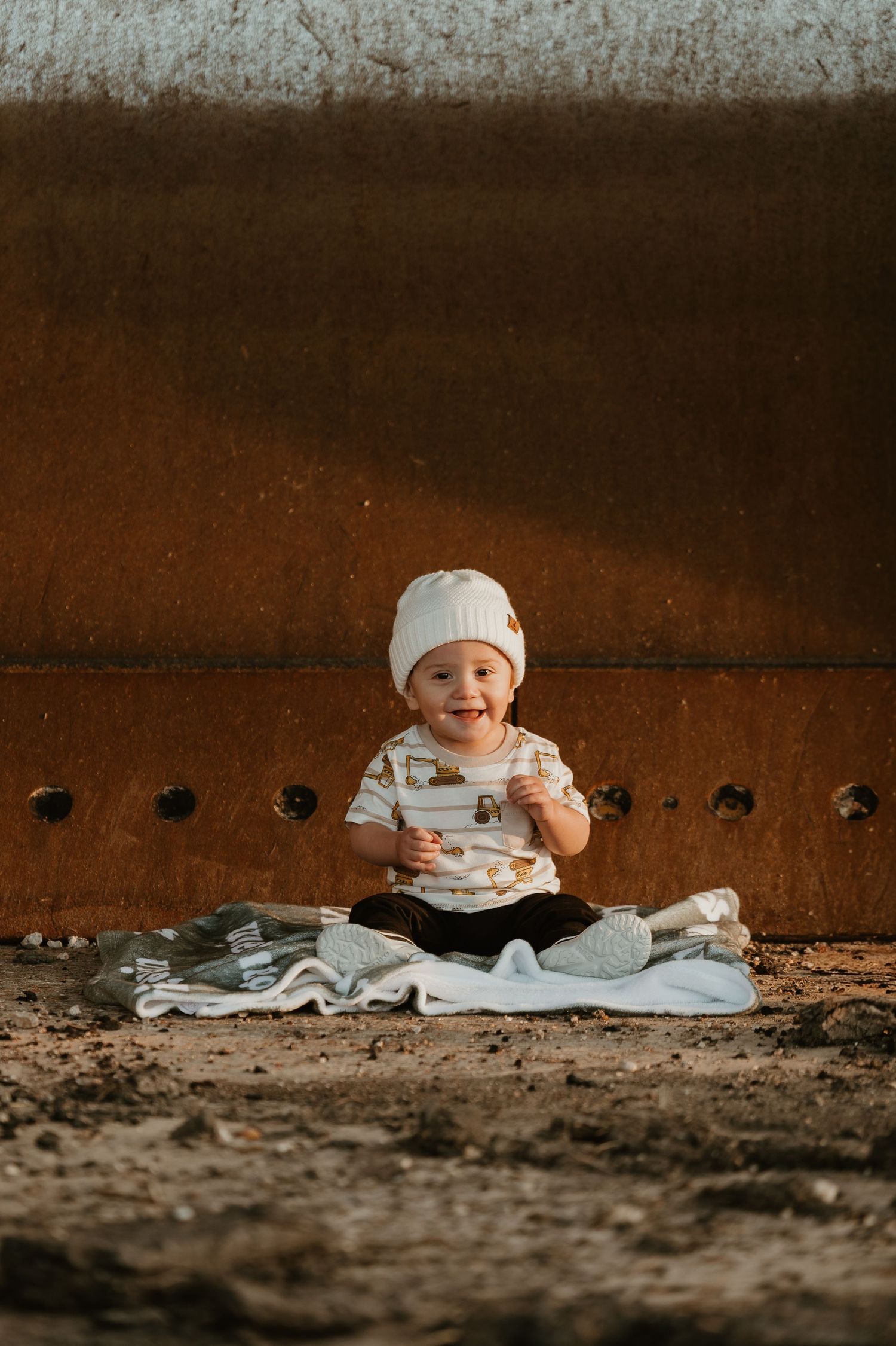 Happy baby in white beanie sits on blanket against rusty metal wall, smiling outdoors in warm golden light.