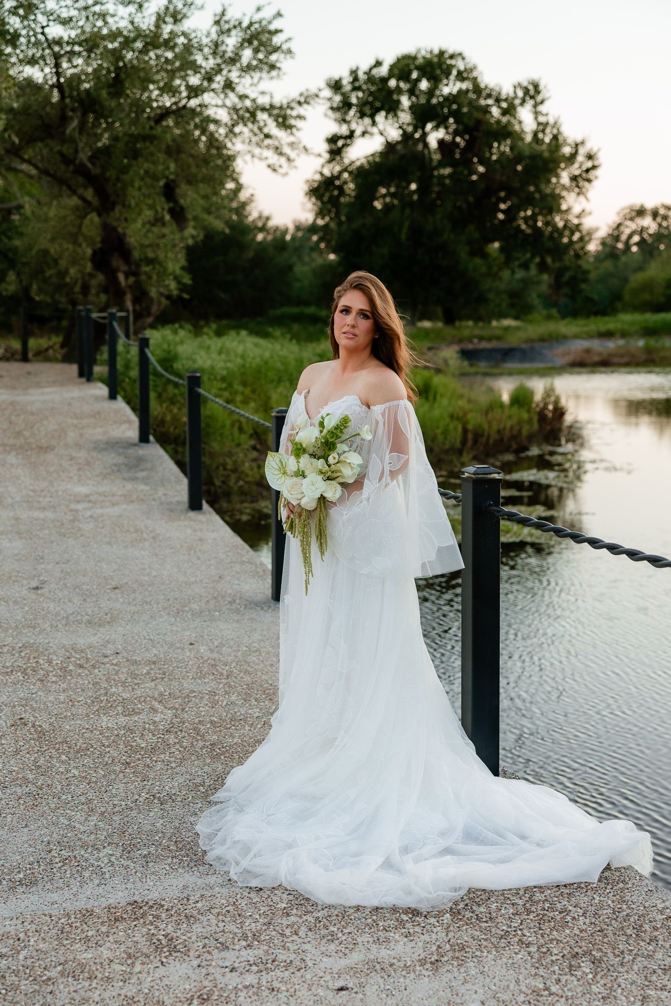 Bride in white off-shoulder tulle gown holding white bouquet on waterfront path surrounded by lush green trees at sunset.