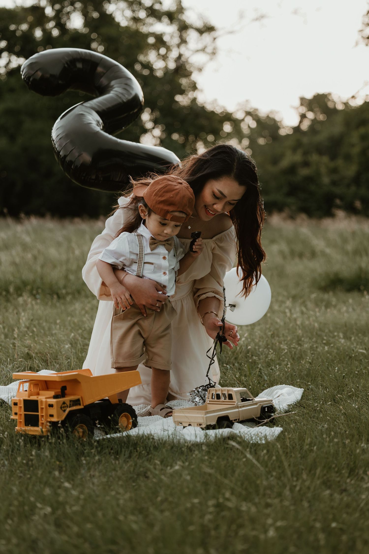 Parent and child playing with toys on picnic blanket under decorative balloons.