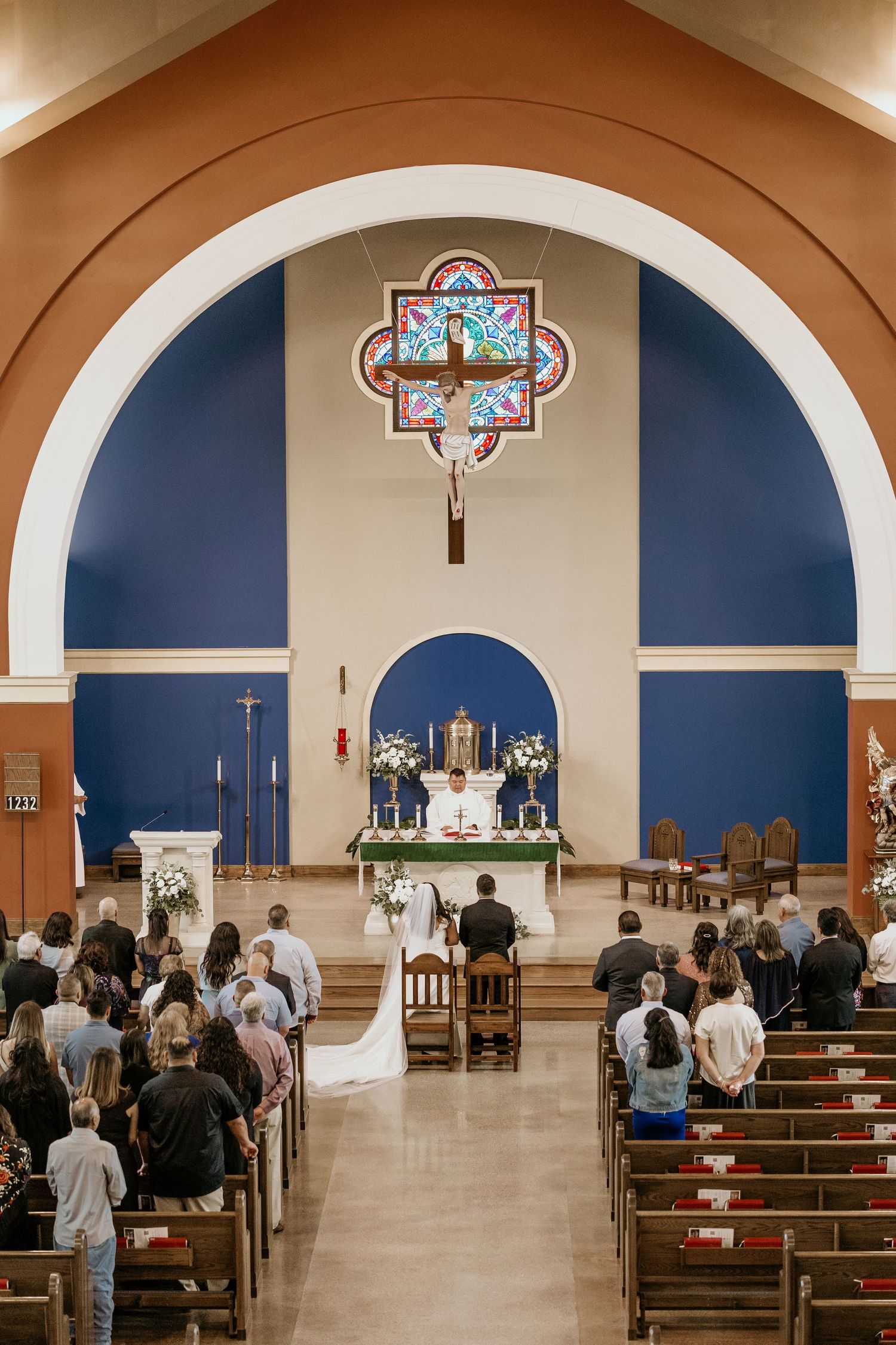Church wedding ceremony with stained glass windows and guests seated in wooden pews.