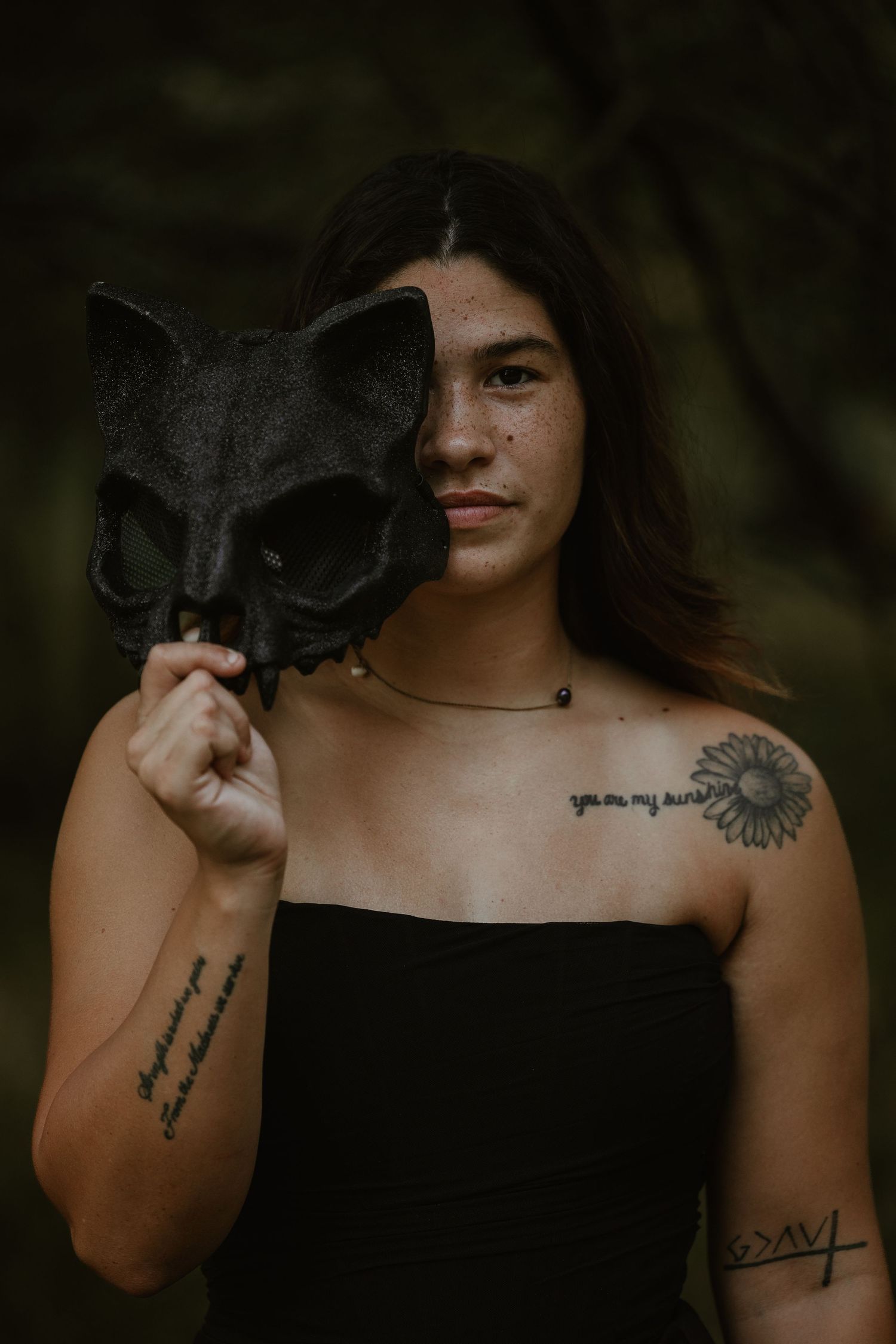 Person in black holding a dark cat mask over half their face with visible tattoos.