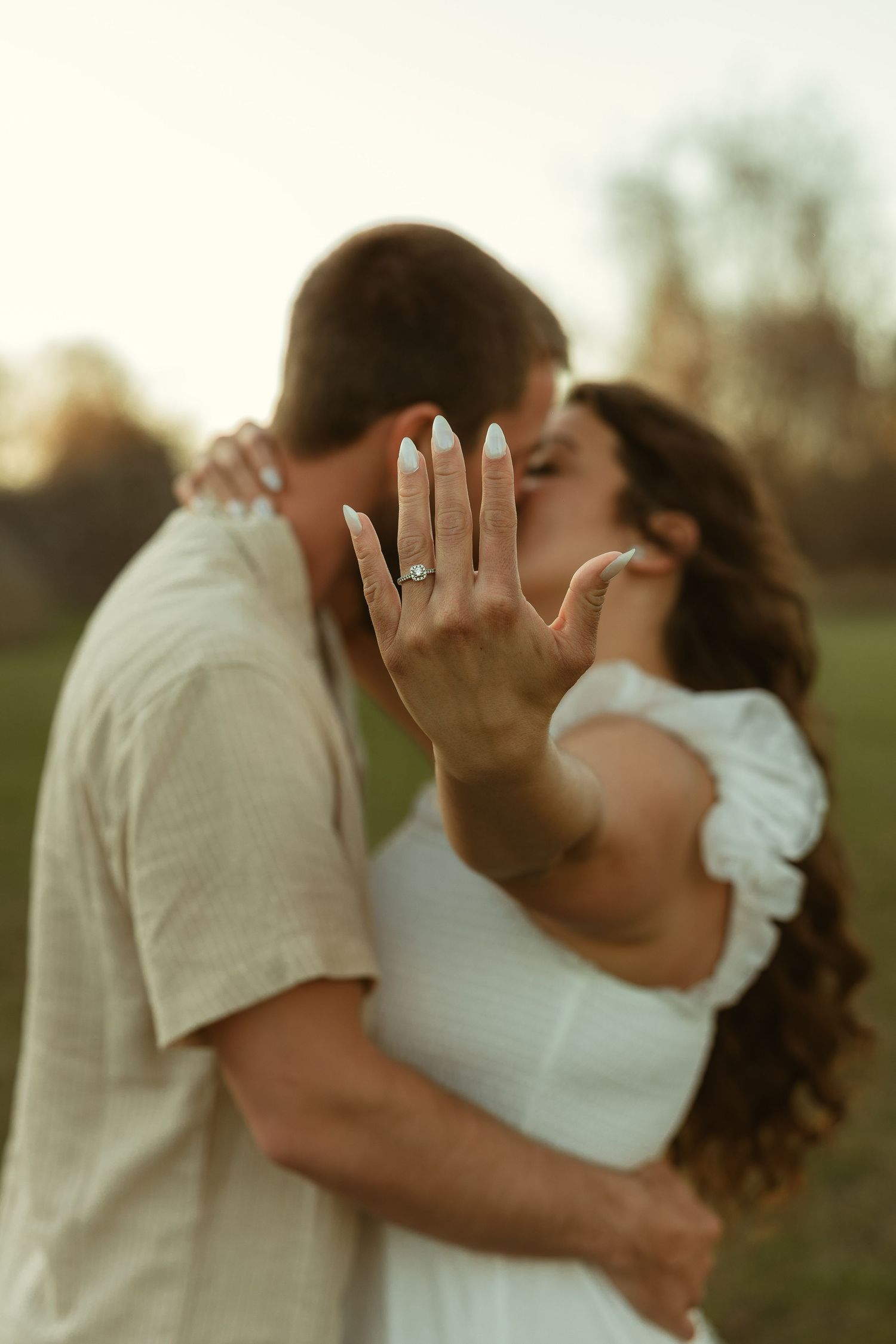 Couple kissing outdoors while showing off an engagement ring, woman in white dress, golden hour background.