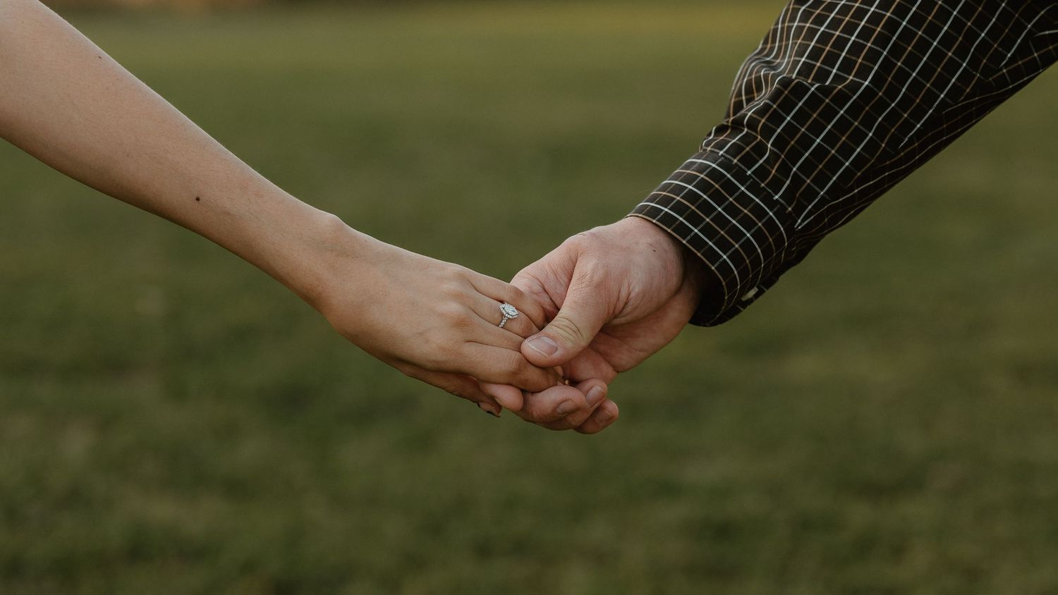Two people holding hands gently outdoors, with a green grass background, symbolizing love and connection.