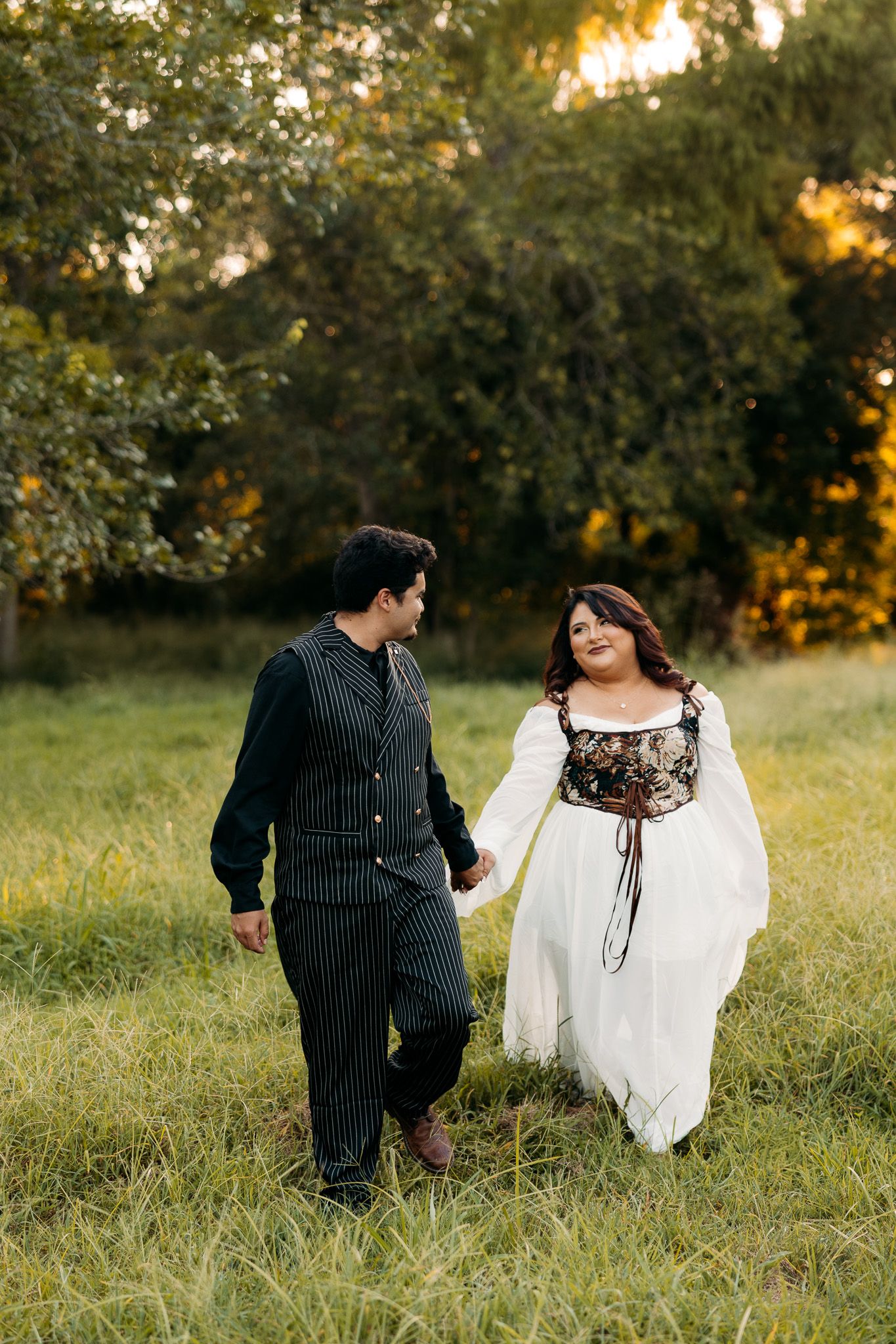 Couple in Renaissance-inspired attire holding hands walking through a lush green field at golden hour.