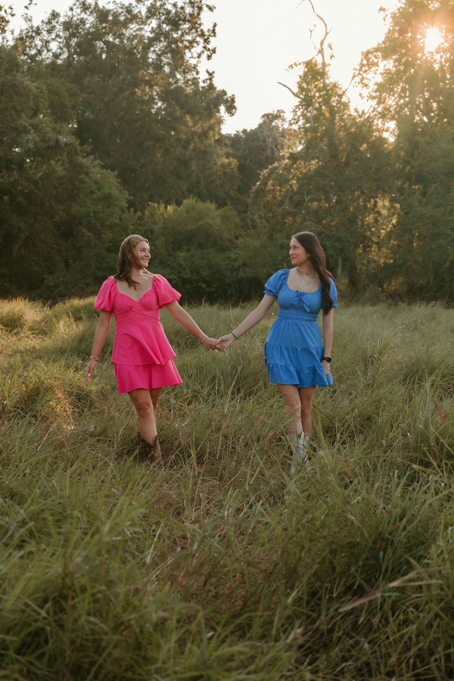 Two girls graduating in colorful summer dresses twirl through tall grass at sunset.