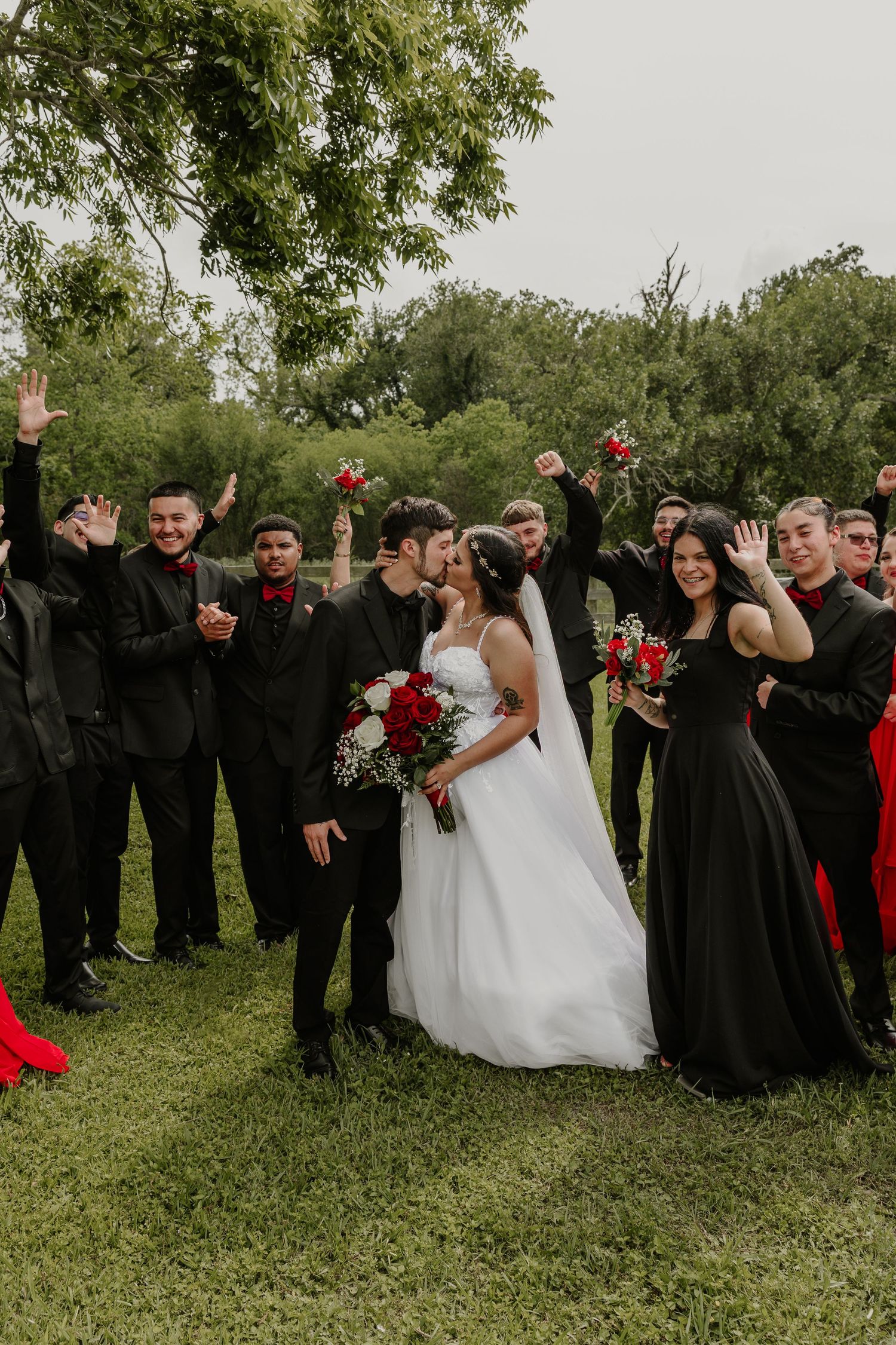 Wedding party in black attire posing playfully on green grass outdoors.