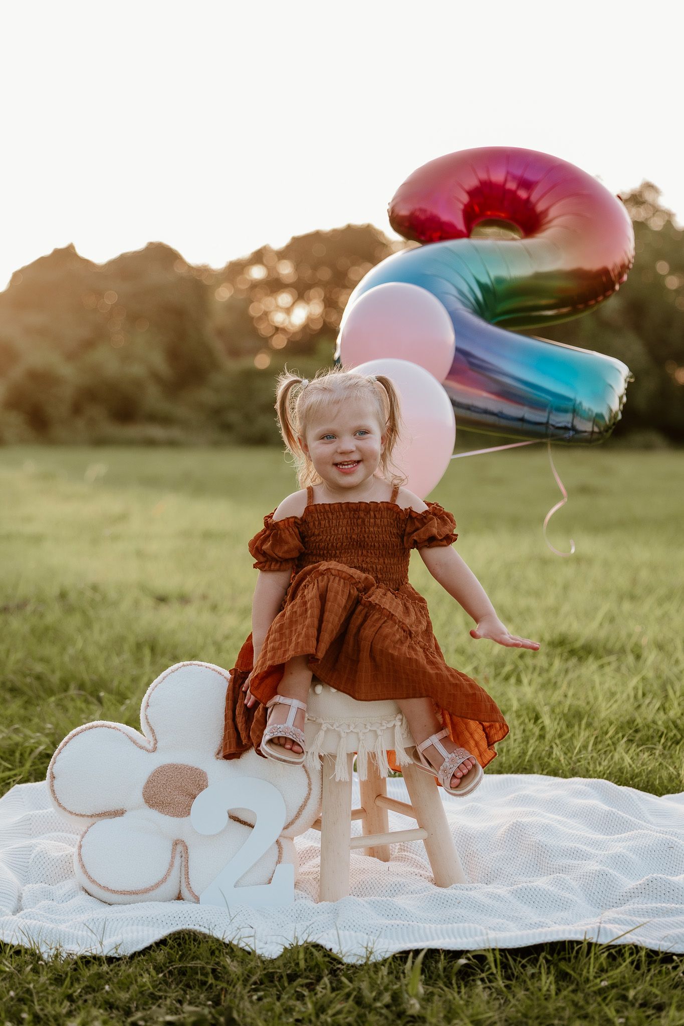 A child in a brown dress stands on a blanket outdoors with colorful number 2 and heart-shaped balloons at sunset.