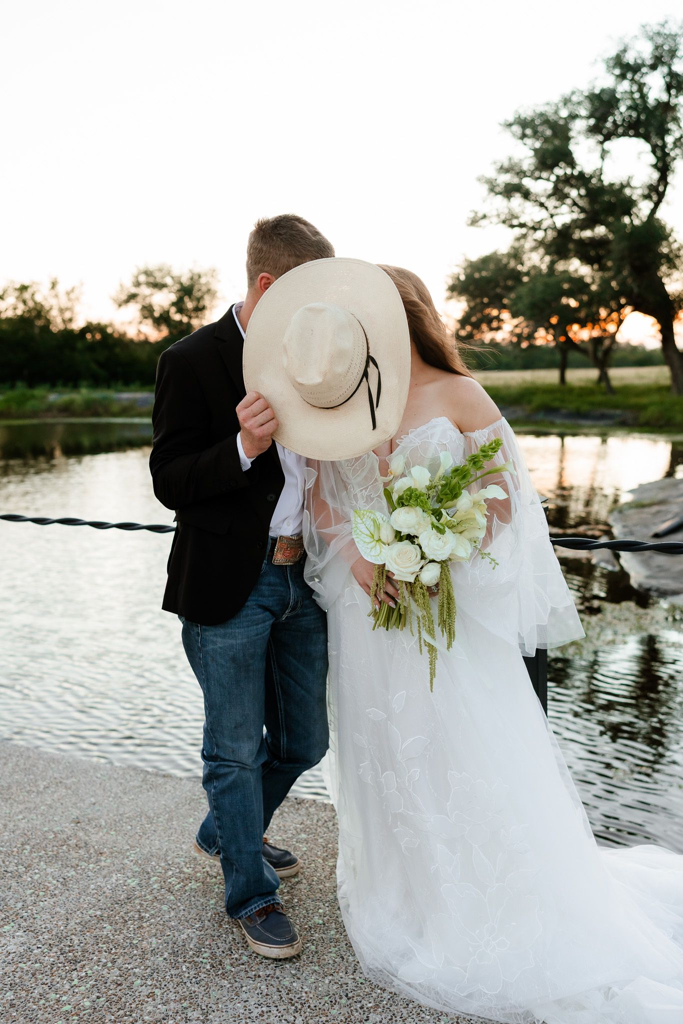Bride and groom share a kiss hidden behind a cowboy hat by a pond at sunset.