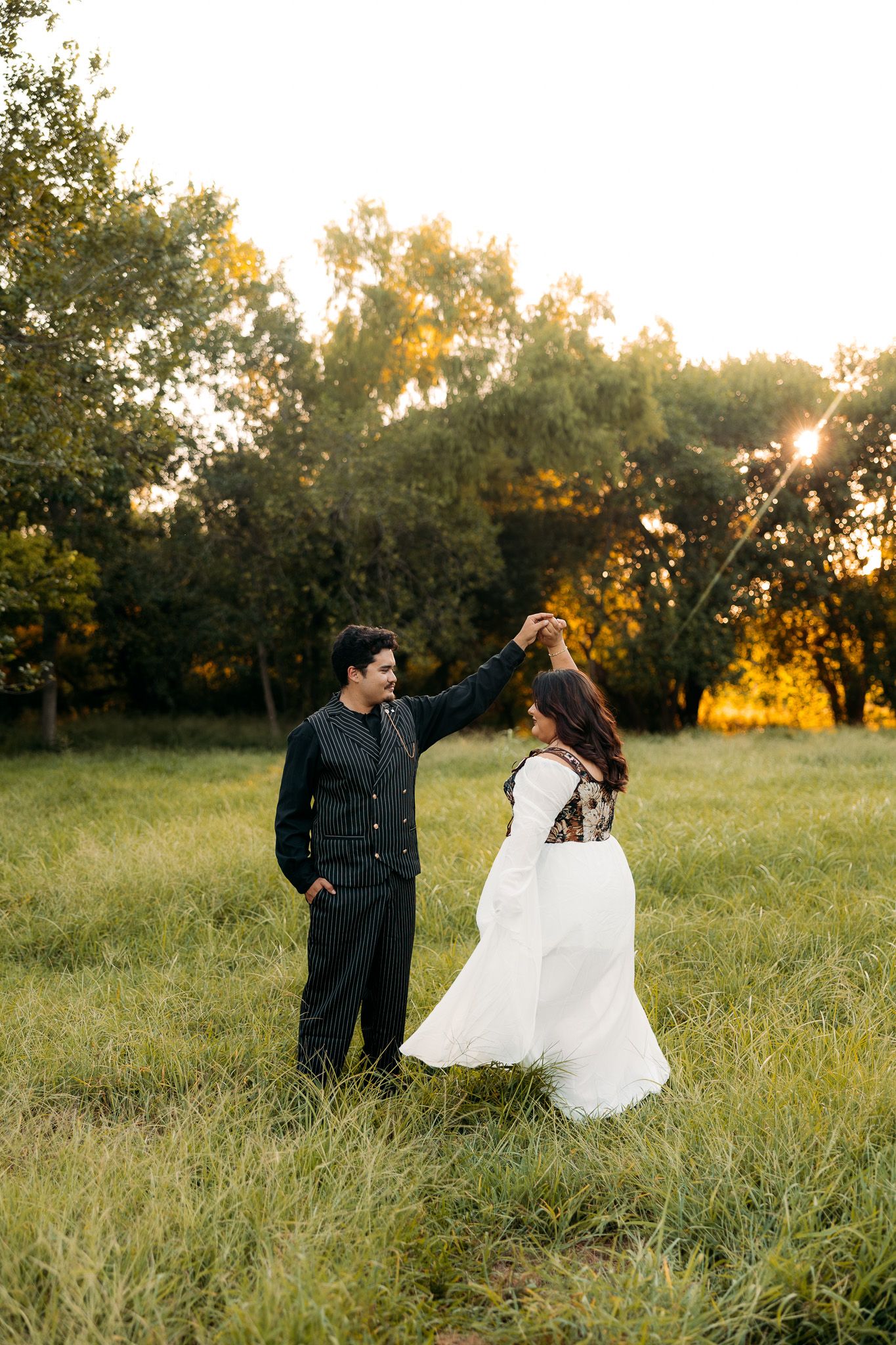 Bride and groom dancing in a sunlit field at golden hour, surrounded by lush green trees.