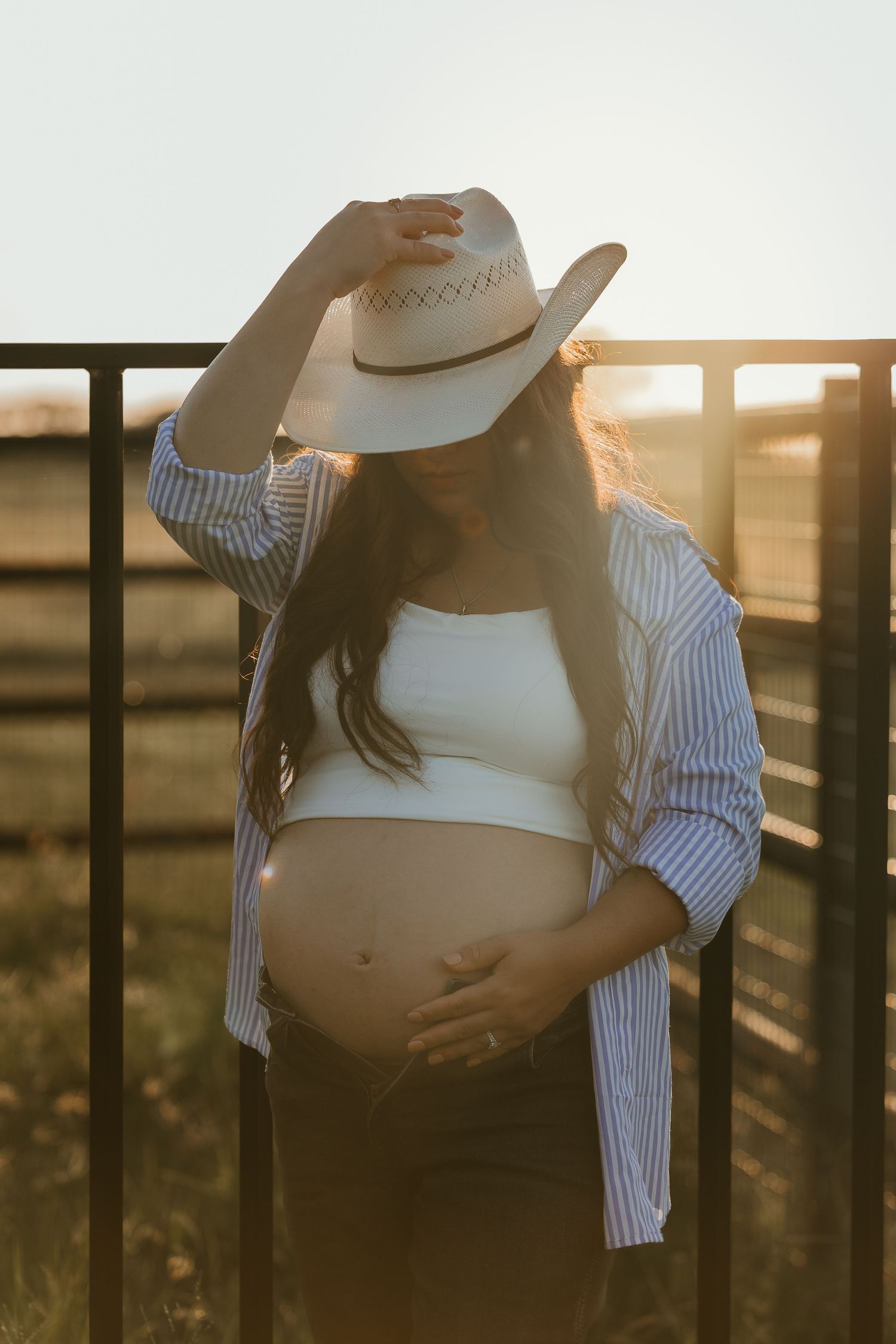 Pregnant woman in white crop top and striped shirt holding baby bump, wearing cowboy hat at golden hour sunset.