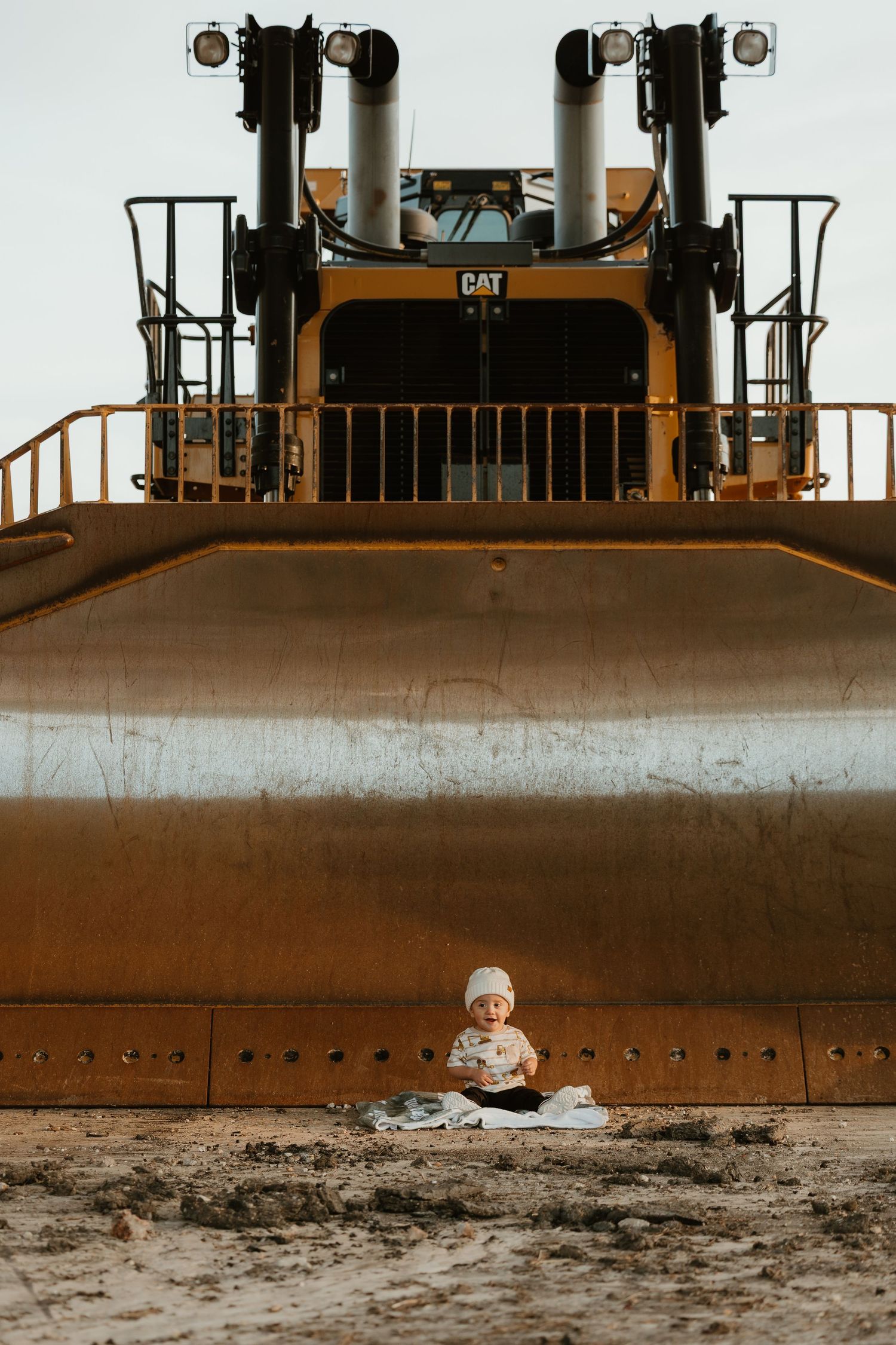 A tiny baby sits in dirt before a massive yellow CAT bulldozer, highlighting the stark size contrast.