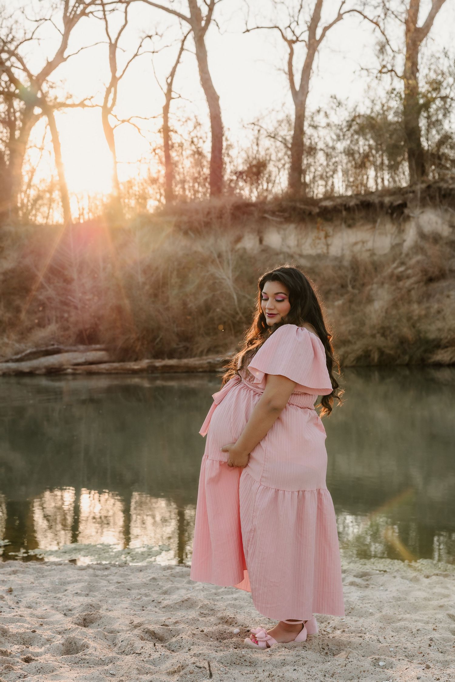 Pregnant woman in pink dress standing by a serene river at golden hour sunset.
