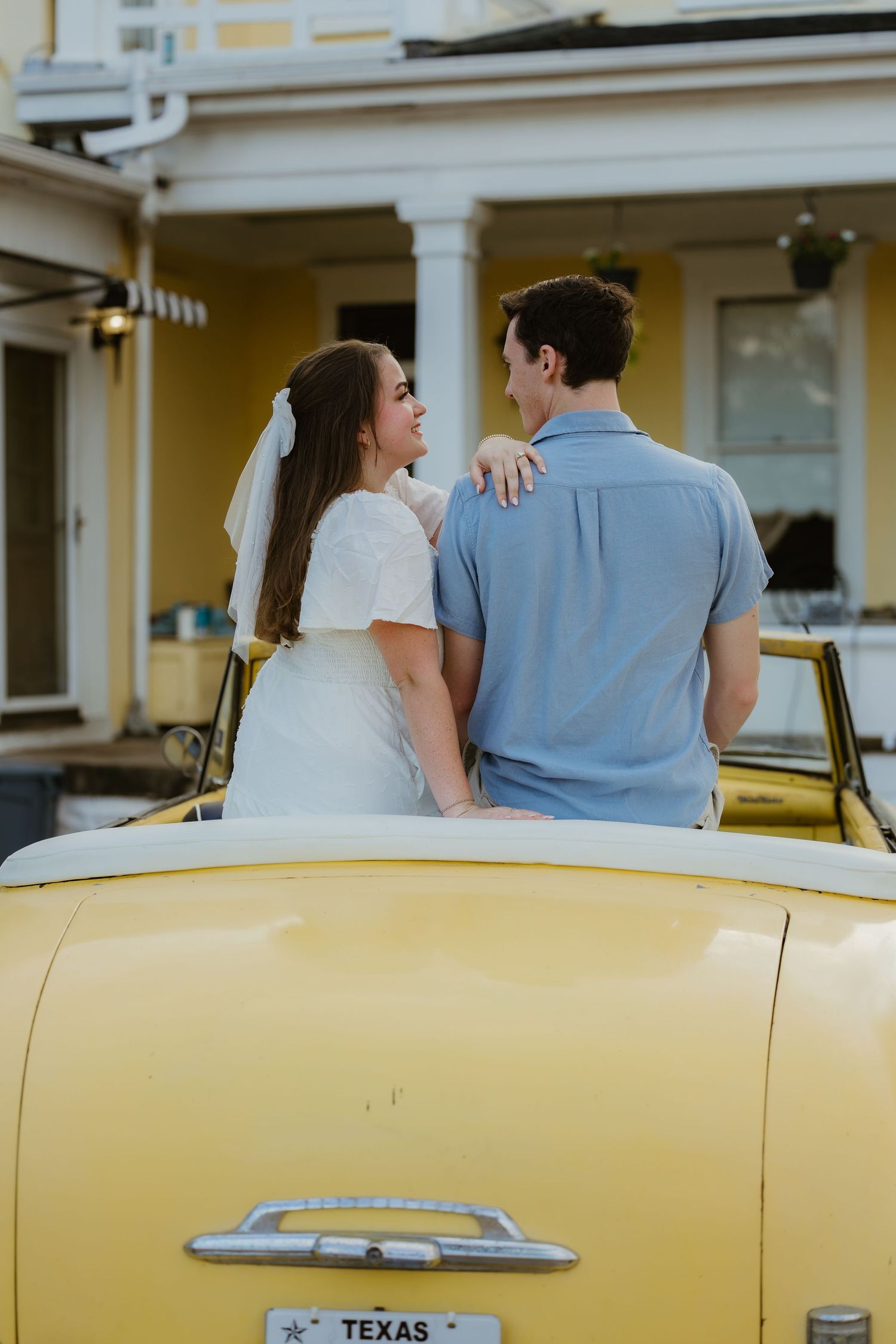 A couple sits on a classic yellow car with Texas plates in front of a yellow building.