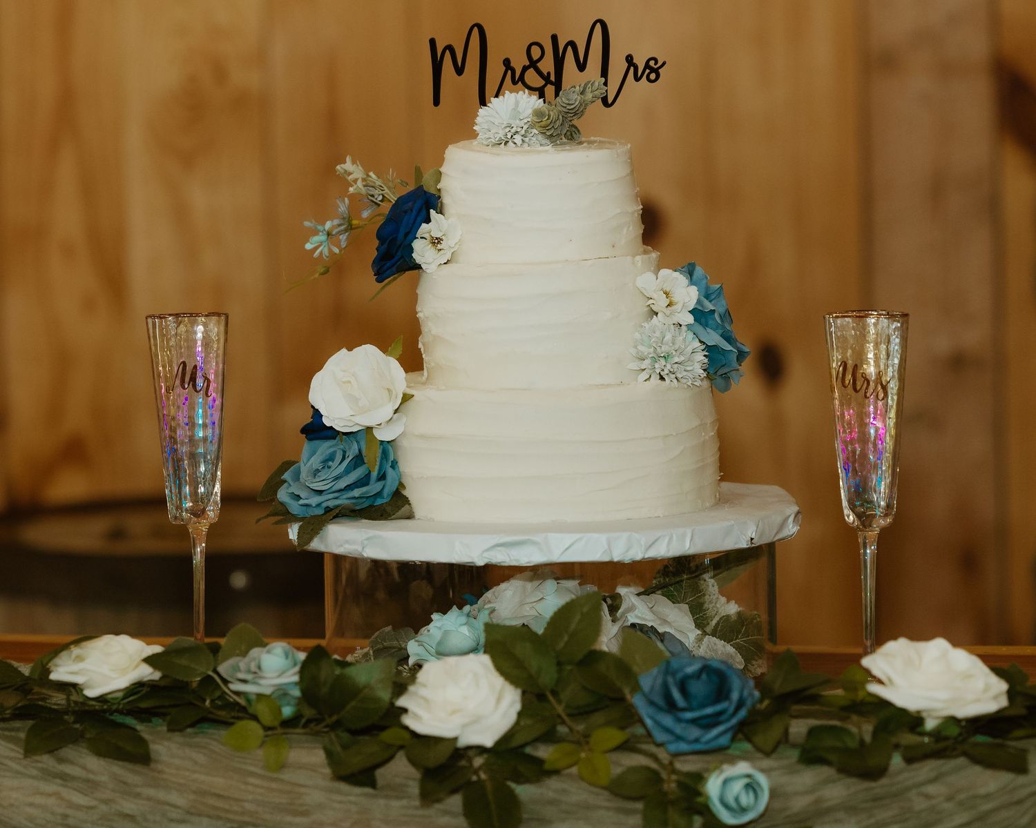 White wedding cake decorated with blue roses and greenery on a rustic wooden table.