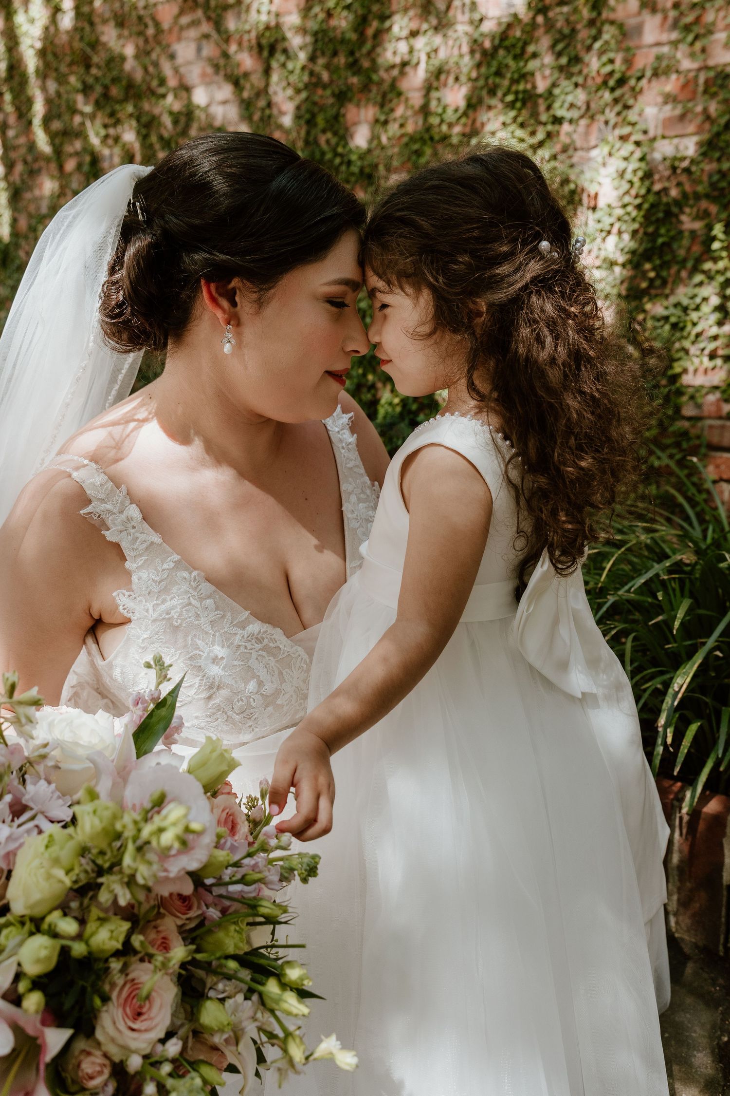 A bride holding a pink bouquet shares an intimate moment against an ivy wall.