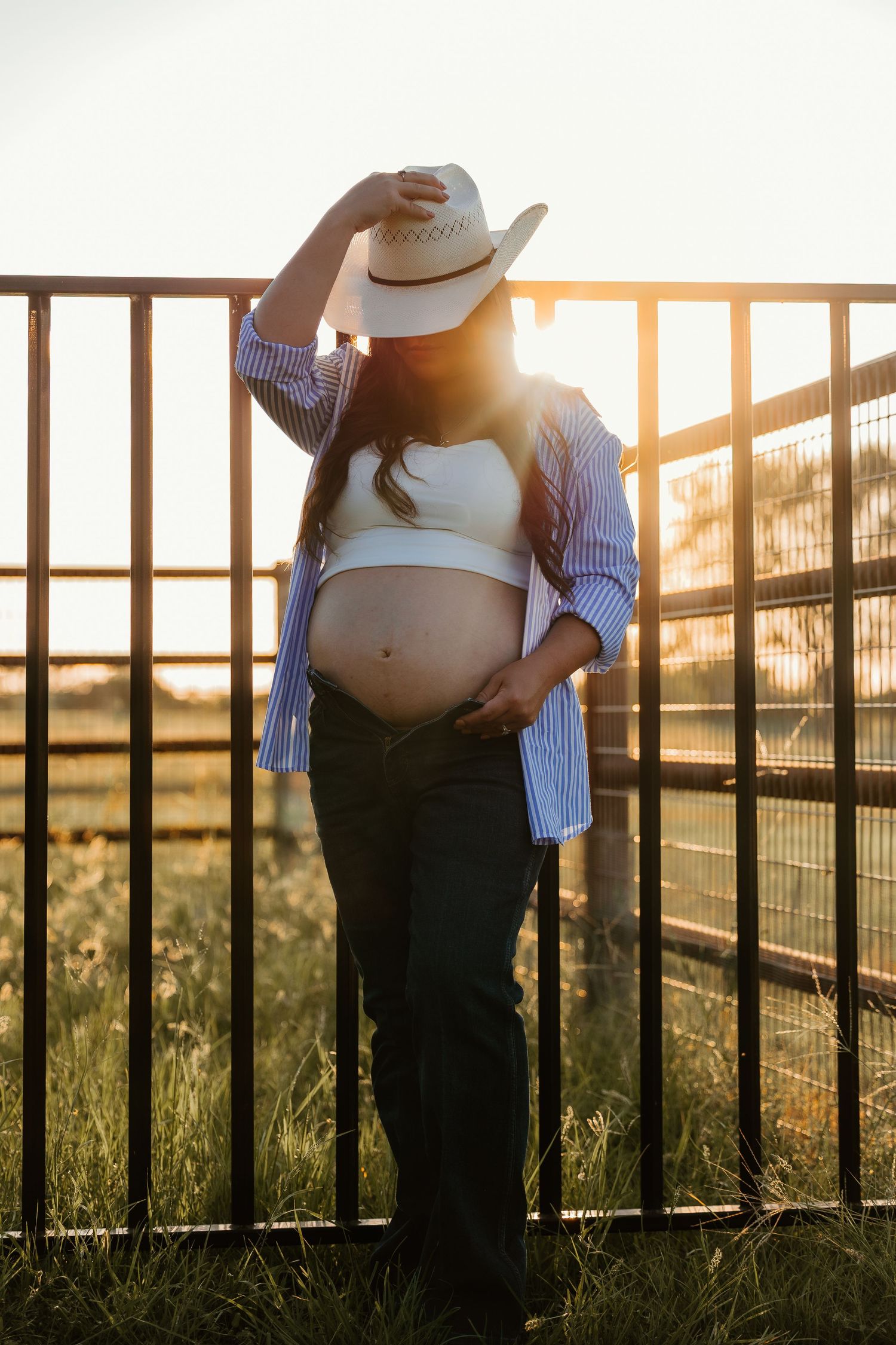 A pregnant woman in western wear poses against a fence during sunset.