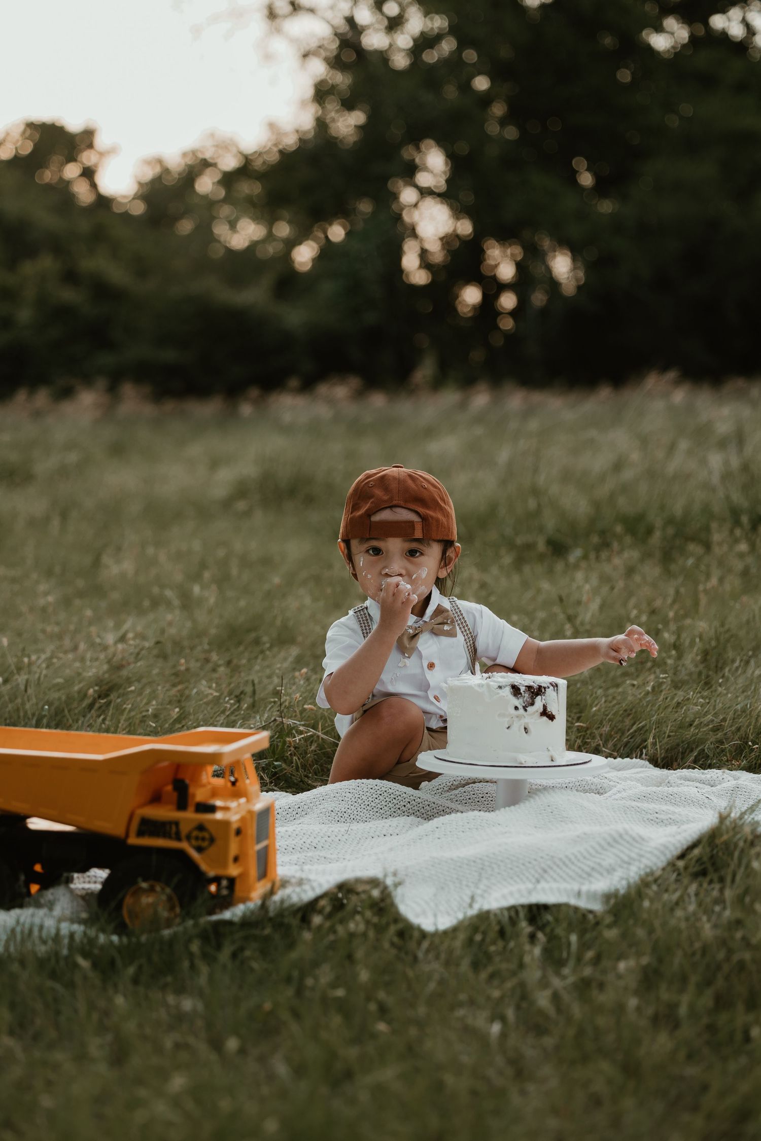 Child in suspenders enjoying cake while sitting on white blanket with toy truck nearby.