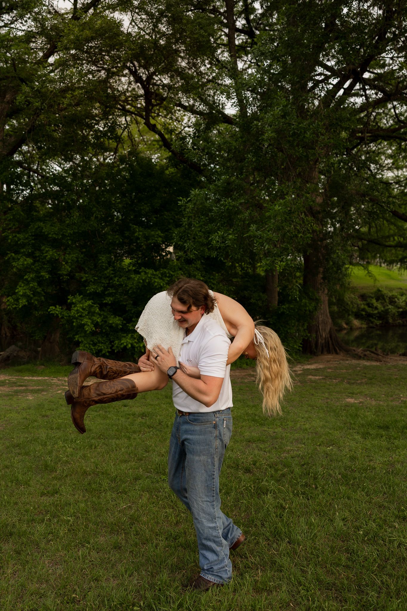 Man in white shirt and jeans lifts woman in wedding dress and cowboy boots outdoors near large green trees.