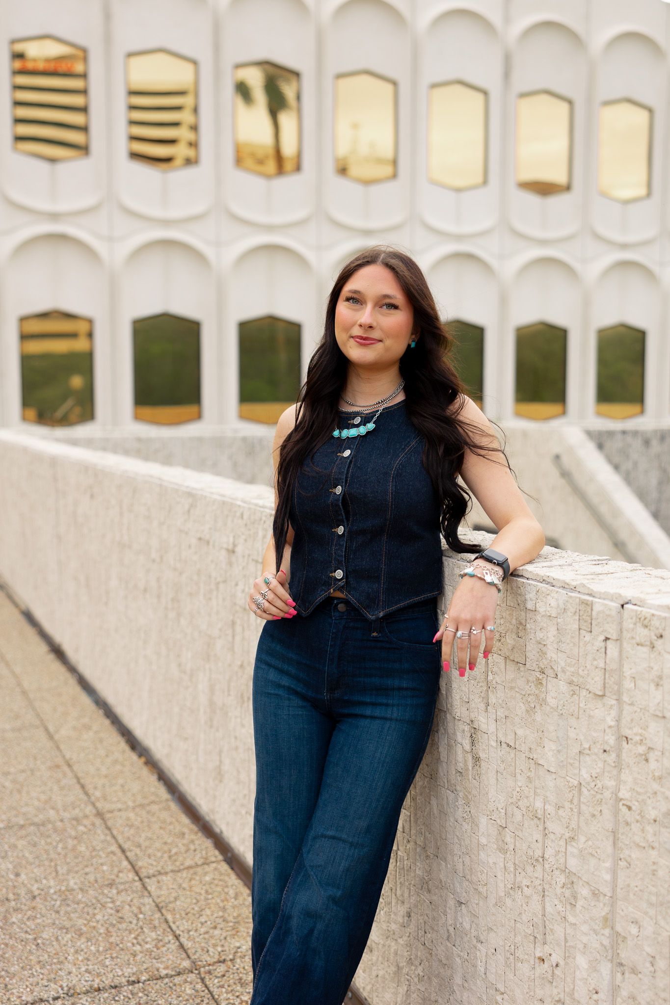 Young woman in denim vest and jeans leaning against a white stone wall outside a modern building.