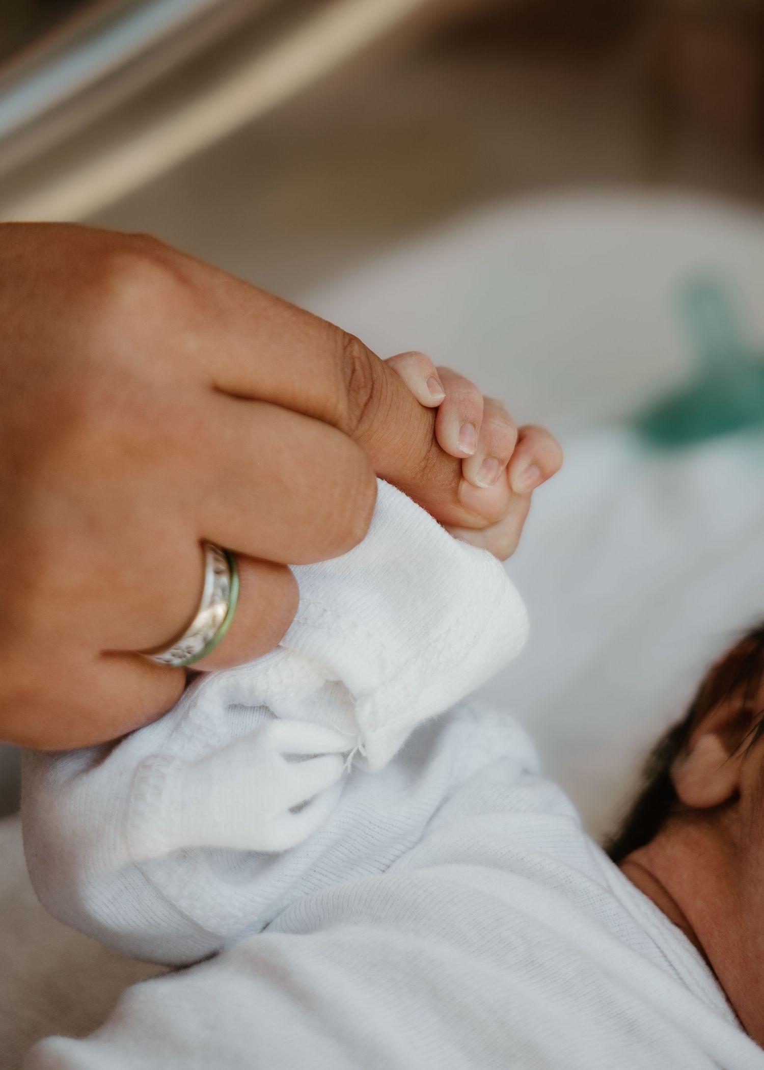 A newborn baby in white clothing gently grips an adult's finger, with a silver ring visible on the parent's hand.