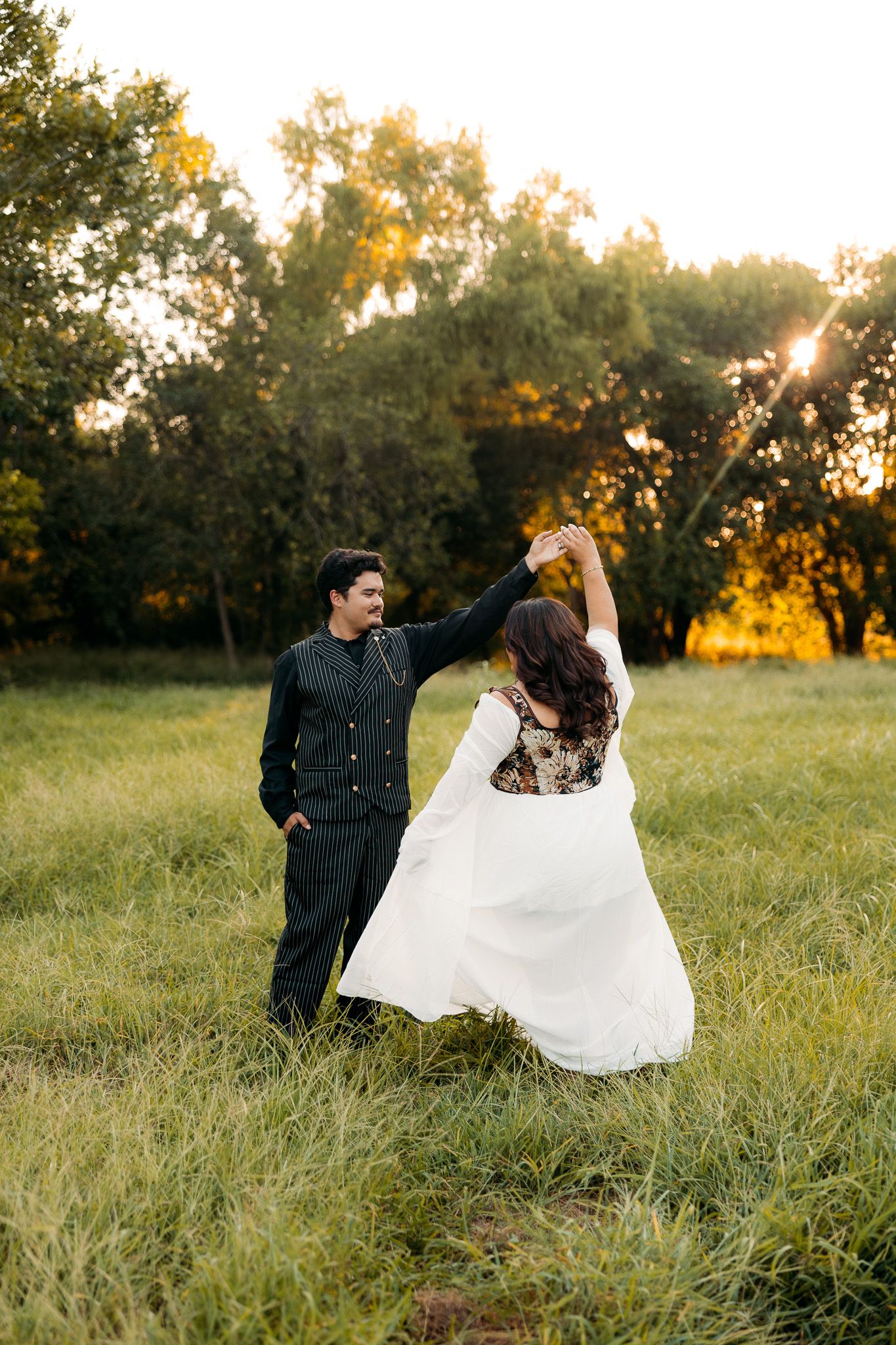 Couple dancing in a sunlit field at golden hour, bride in white gown twirling as groom smiles in black suit.